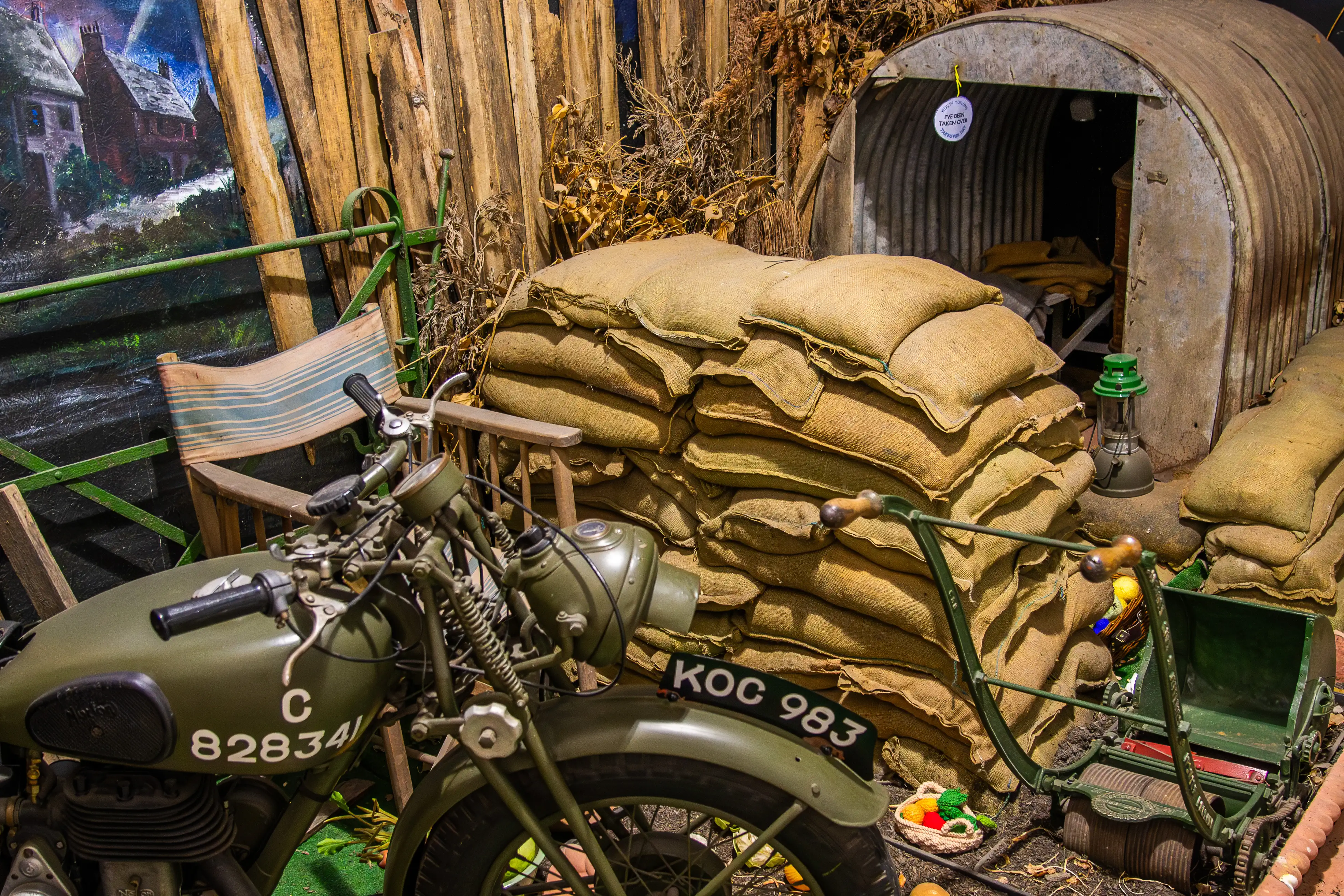 A vintage military motorcycle with the markings 'KOC 983' and 'C 828341' parked in front of a makeshift shelter made of sandbags. A striped deck chair, a green lantern, and a wooden fence are visible, with a painted rural house in the background