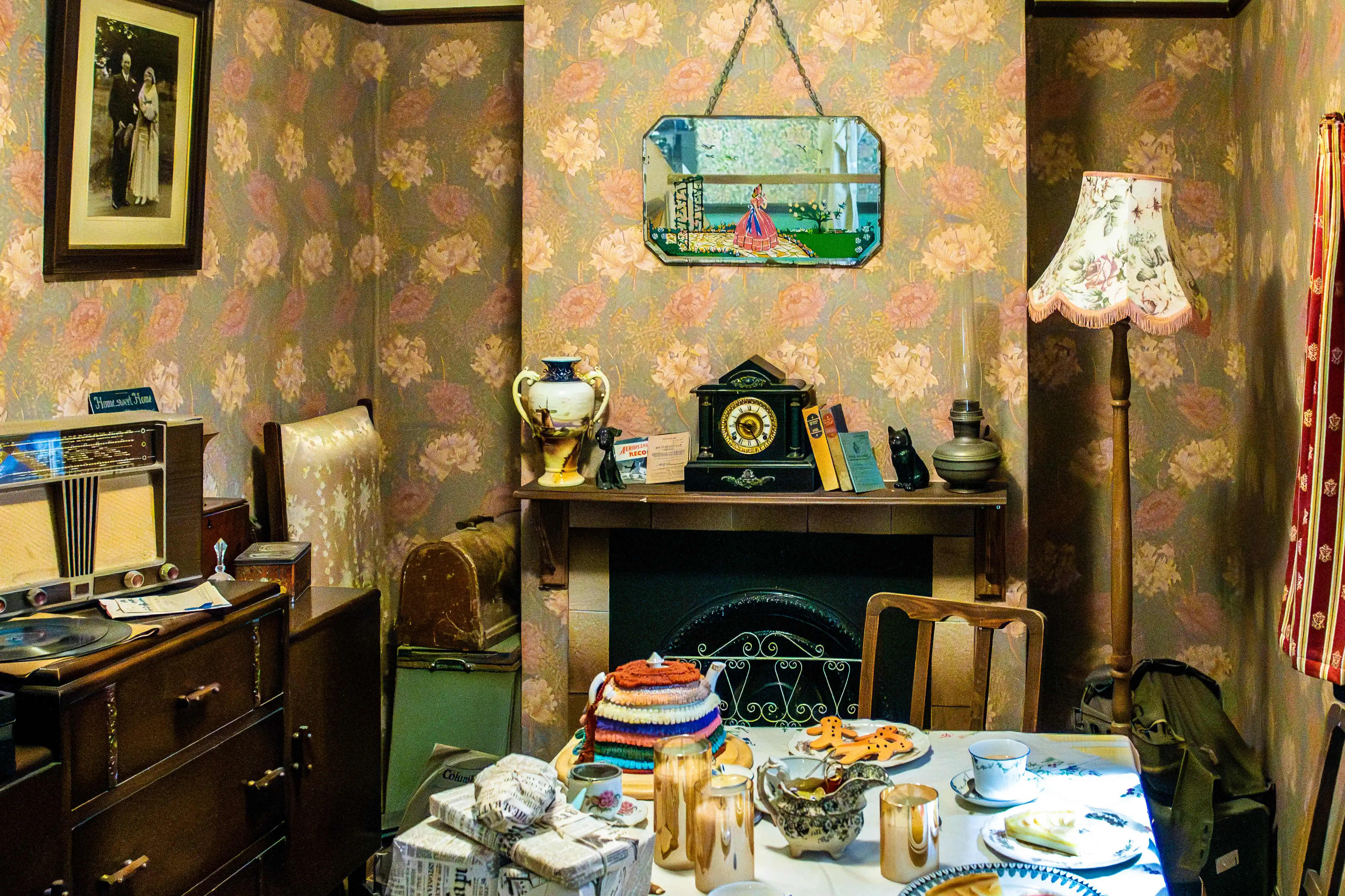 This image presents a meticulously recreated or preserved vintage living room, likely from the 1950s. The walls are covered in a distinctive floral wallpaper featuring a pattern of large, faded pink and yellow flowers against a muted grey-green background. To the left, a framed sepia-toned photograph of a formal couple hangs on the wall above a vintage wooden dresser. The dresser supports a classic 1950s radio with glowing indicators and a turntable, alongside a few vinyl records. A chair with a patterned fabric is partially visible behind the dresser. The focal point of the room is a fireplace, adorned with a dark mantelpiece. On the mantel sits a decorative urn, a small black figurine of a dog, a 'Aeroplane Record' pamphlet, a classic black mantel clock with a gold face, two books, and another black cat figurine. Above the mantelpiece, an octagonal mirror with a bevelled edge hangs from a chain, reflecting a scene of a woman in a pink dress in a garden. In front of the fireplace, a small table is set for tea. It features a teapot covered in a colourful knitted cosy, a delicate teacup and saucer, a plate with ginger-nut biscuits shaped like animals, and a slice of cake on another plate. A decorative teapot or gravy boat and several metallic candle holders are also present. To the left of the table, several packages wrapped in newspaper and tied with string are stacked. To the right of the fireplace, a tall wooden floor lamp with a fringed floral lampshade stands. A vintage kerosene lamp sits on a side table next to it. A striped curtain hangs on the far right wall. The overall impression is one of a well-lived-in, comforting space, rich with personal items and the aesthetic of a bygone era.