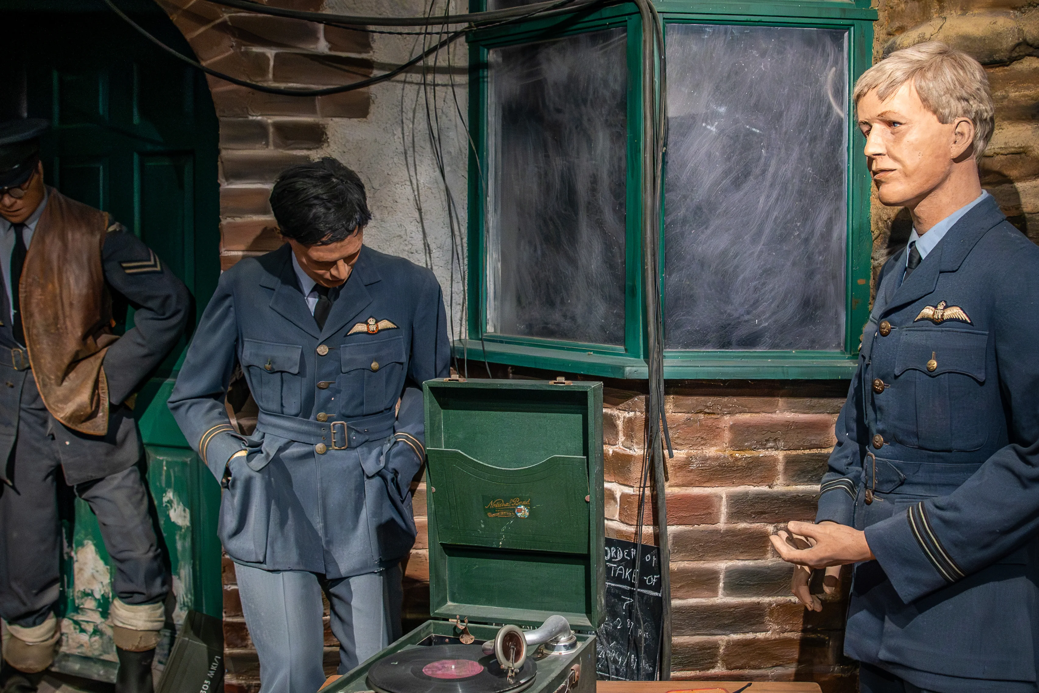 Close scene from a 1940s house display: uniformed staff beside a green record player cabinet in front of a brick wall and a green-framed window.  