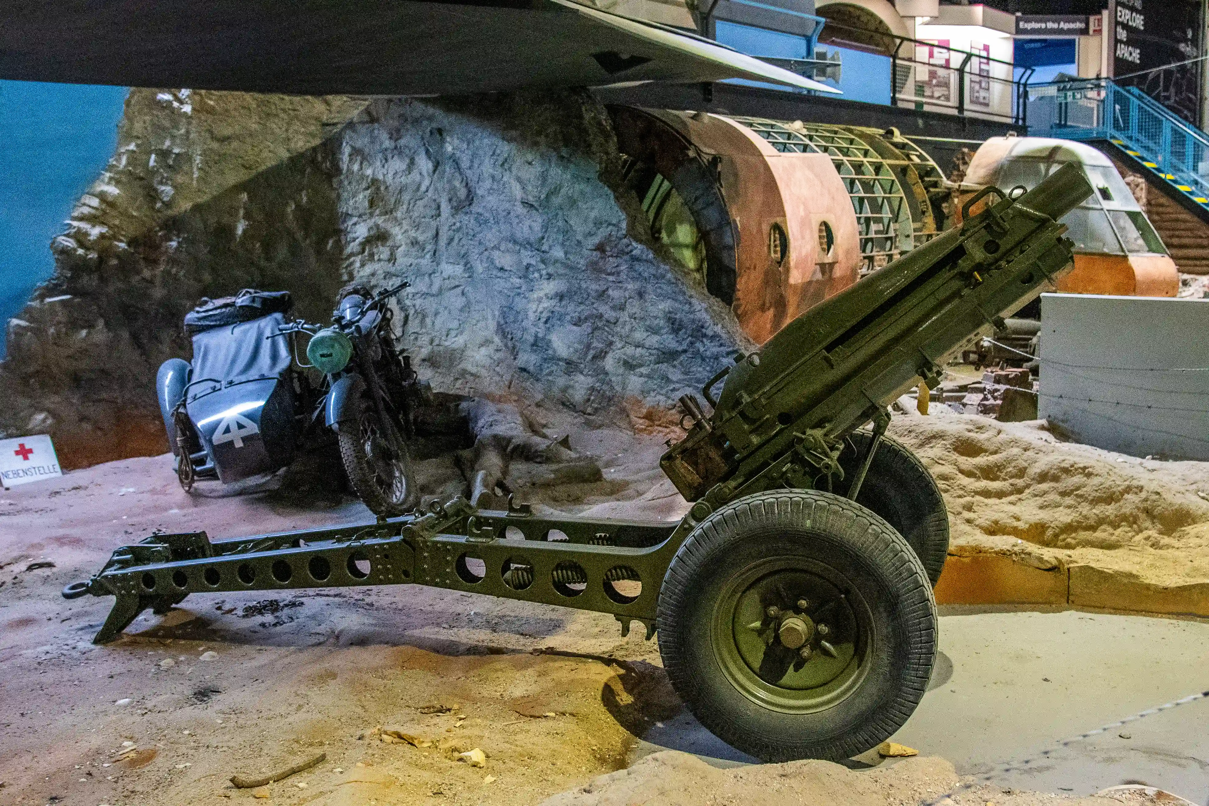 A green artillery gun is positioned in the foreground of a museum exhibit. Behind it, a dark blue motorcycle with a sidecar is parked. In the background, a rocky display and part of an aircraft fuselage are visible.