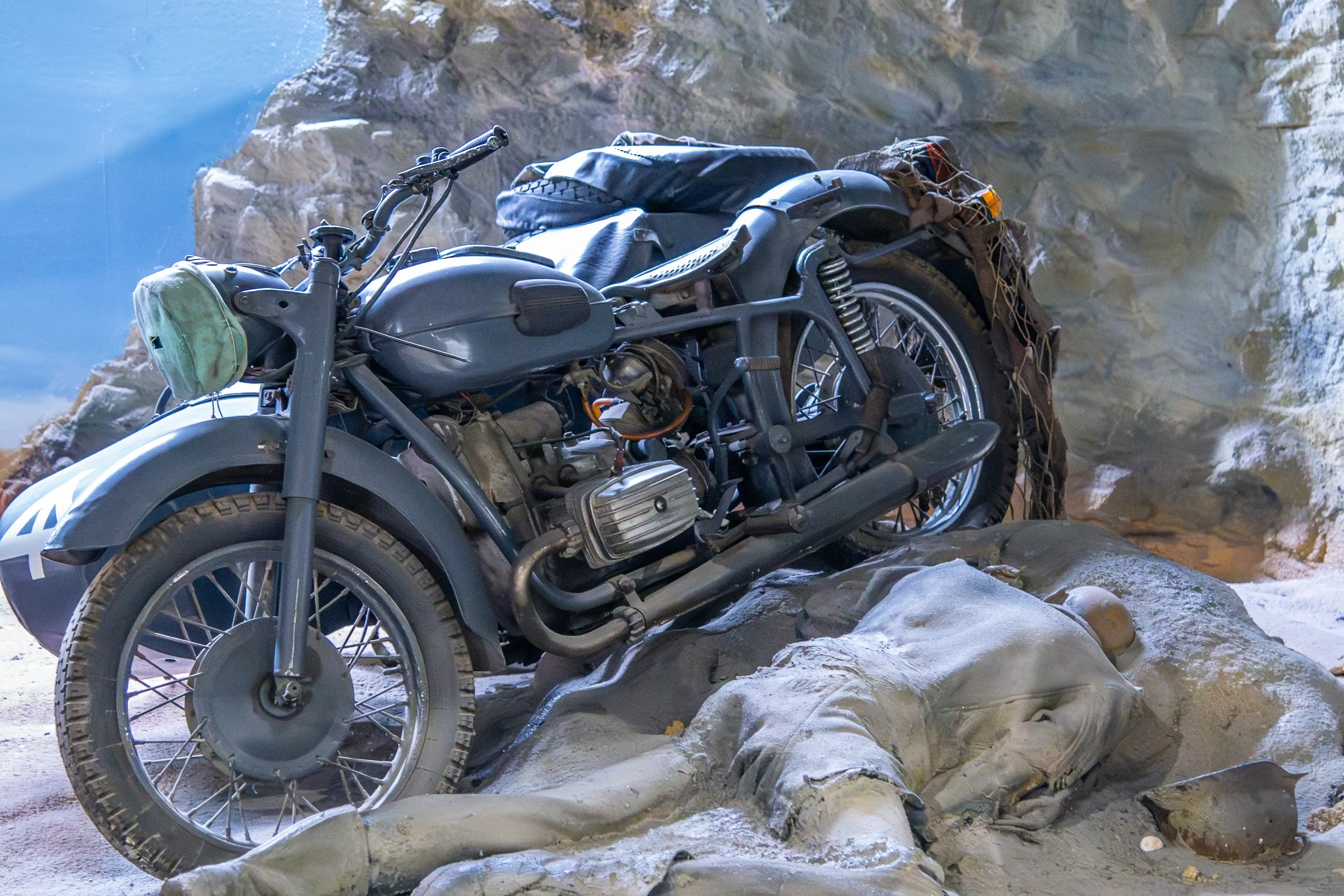 A grey military motorcycle is parked beside a mannequin of a fallen soldier dressed in wartime clothing, lying on a sandy, rocky surface, with a painted rocky background.