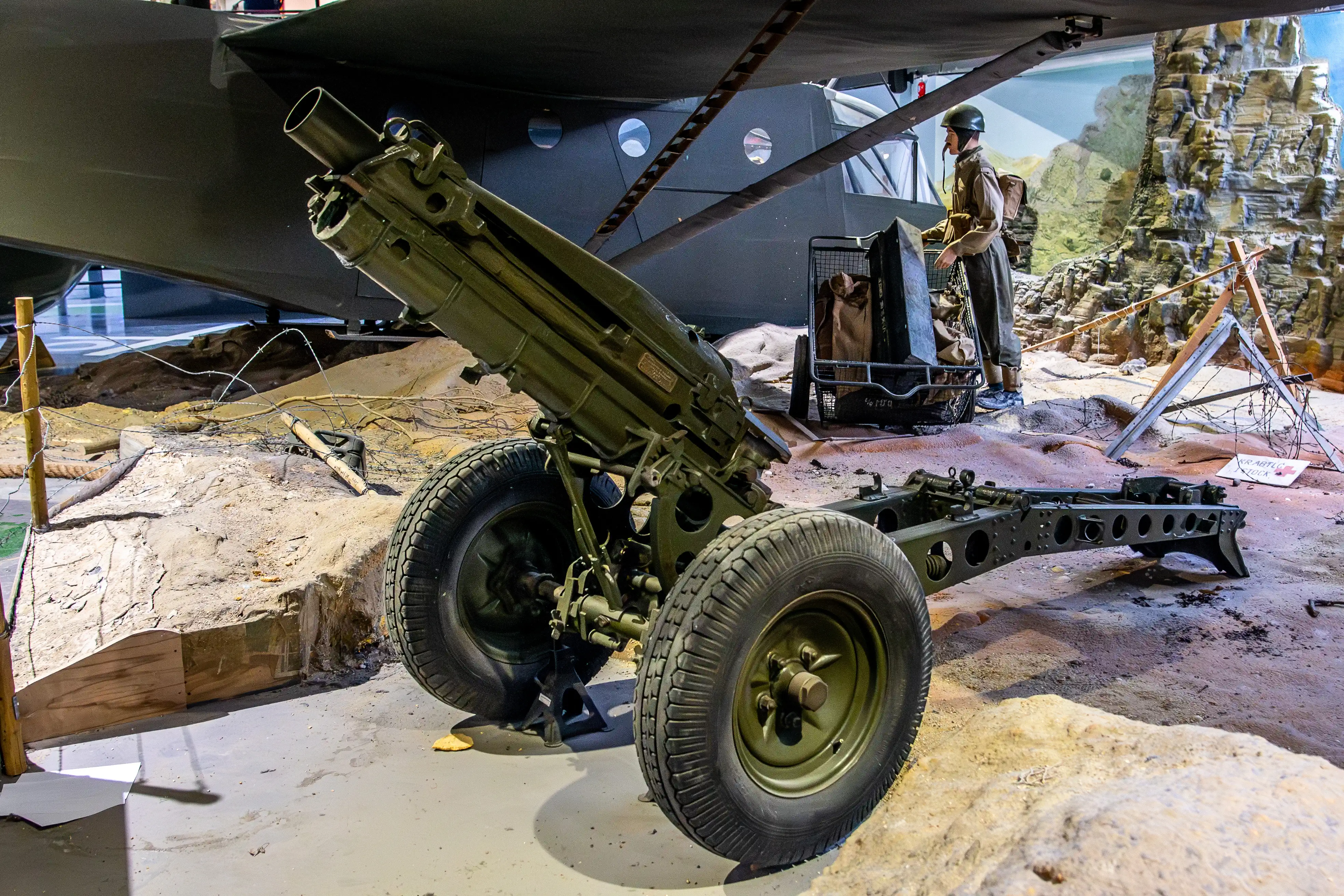 A military artillery gun with large wheels is displayed in a museum exhibit, angled towards the viewer. In the background, part of a dark-coloured aircraft is visible, along with a mannequin in a military uniform standing near a cart and a rocky landscape.