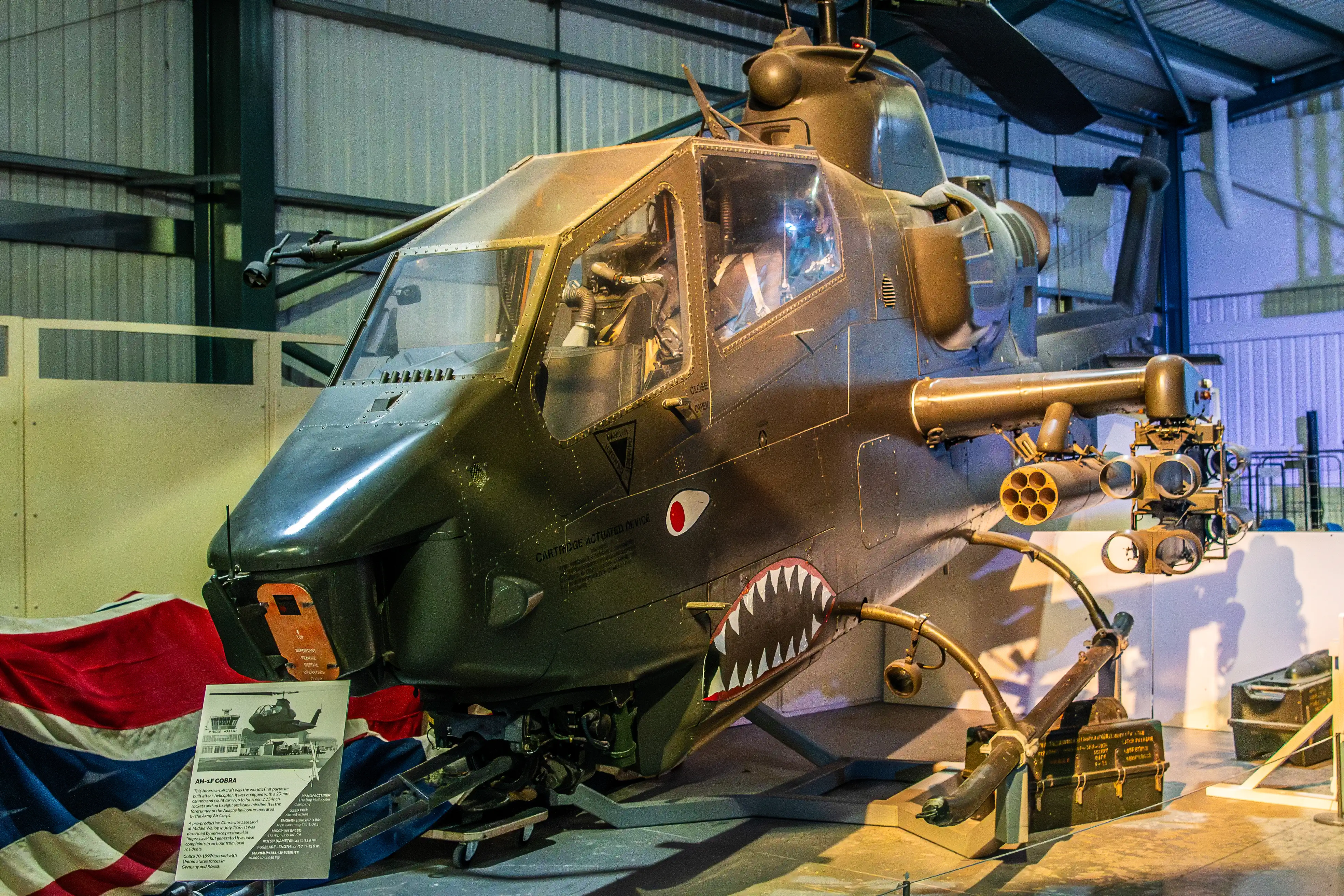 A dark green AH-1S Cobra attack helicopter with a shark-mouth graphic and weaponry is parked indoors, with a Union Jack flag to its left and an information sign in the foreground.