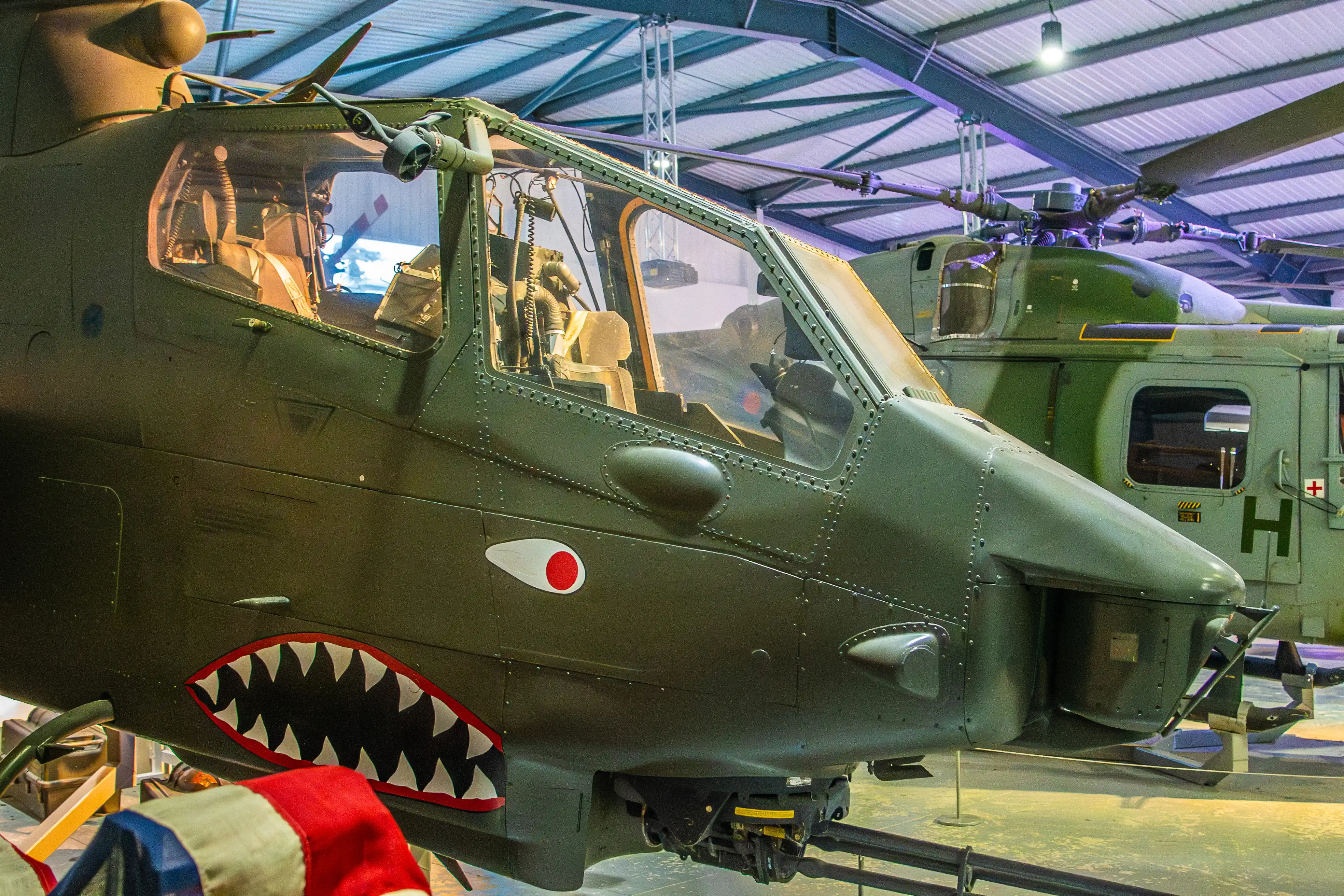 Close-up of the nose of a green military attack helicopter with a large, black shark mouth painted on its side, a white-outlined red circle insignia, and a glimpse into the cockpit. A second helicopter is behind it.