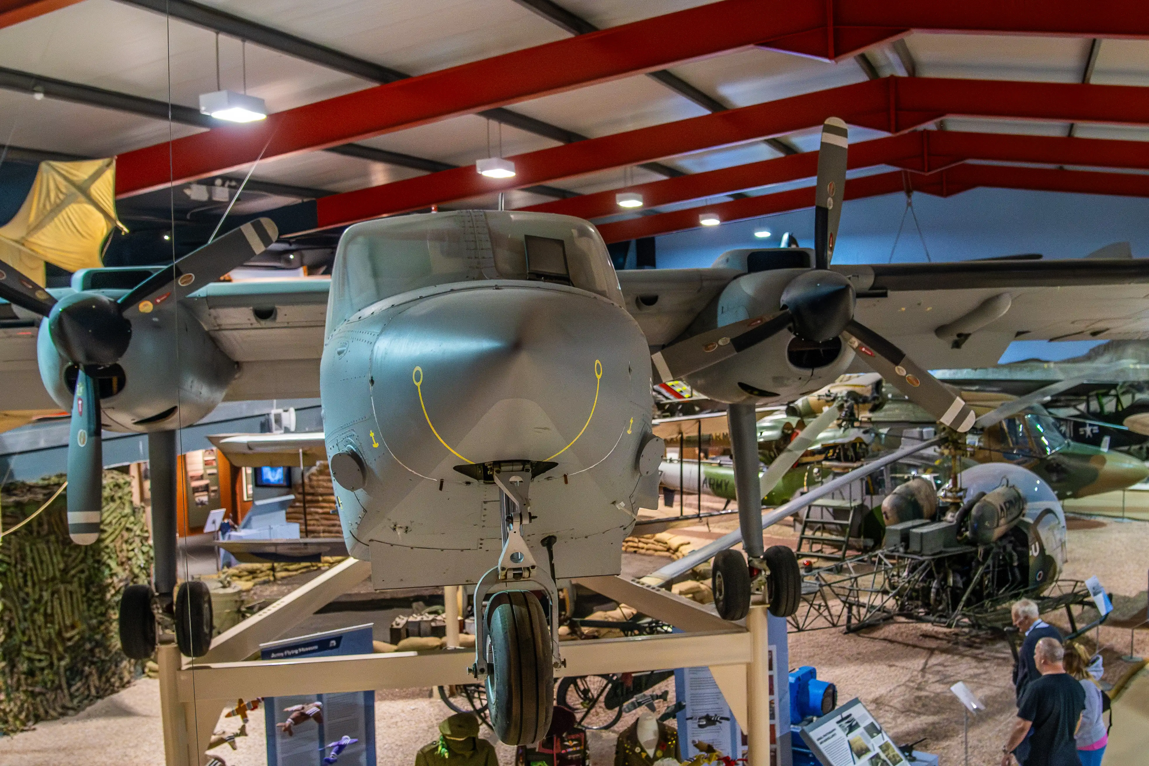 A grey twin-engine propeller aircraft is displayed prominently in a museum. Its nose is facing the camera and has a yellow marking. Other aircraft and visitors are visible in the background within the hangar.