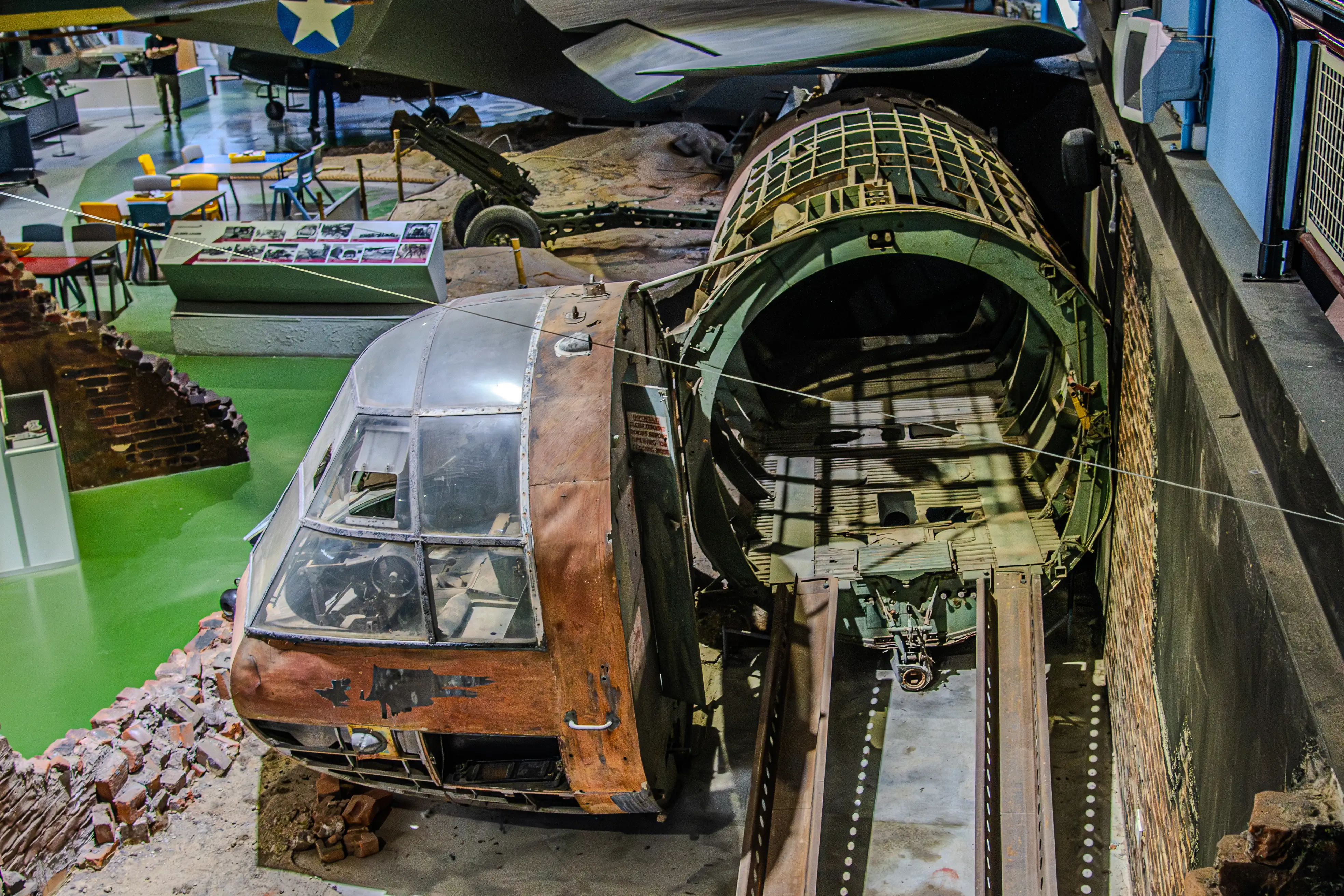 Overhead view of a museum exhibit featuring the front cockpit section and a portion of the hollow fuselage of a large aircraft, presented as if damaged and in a simulated ruin environment.