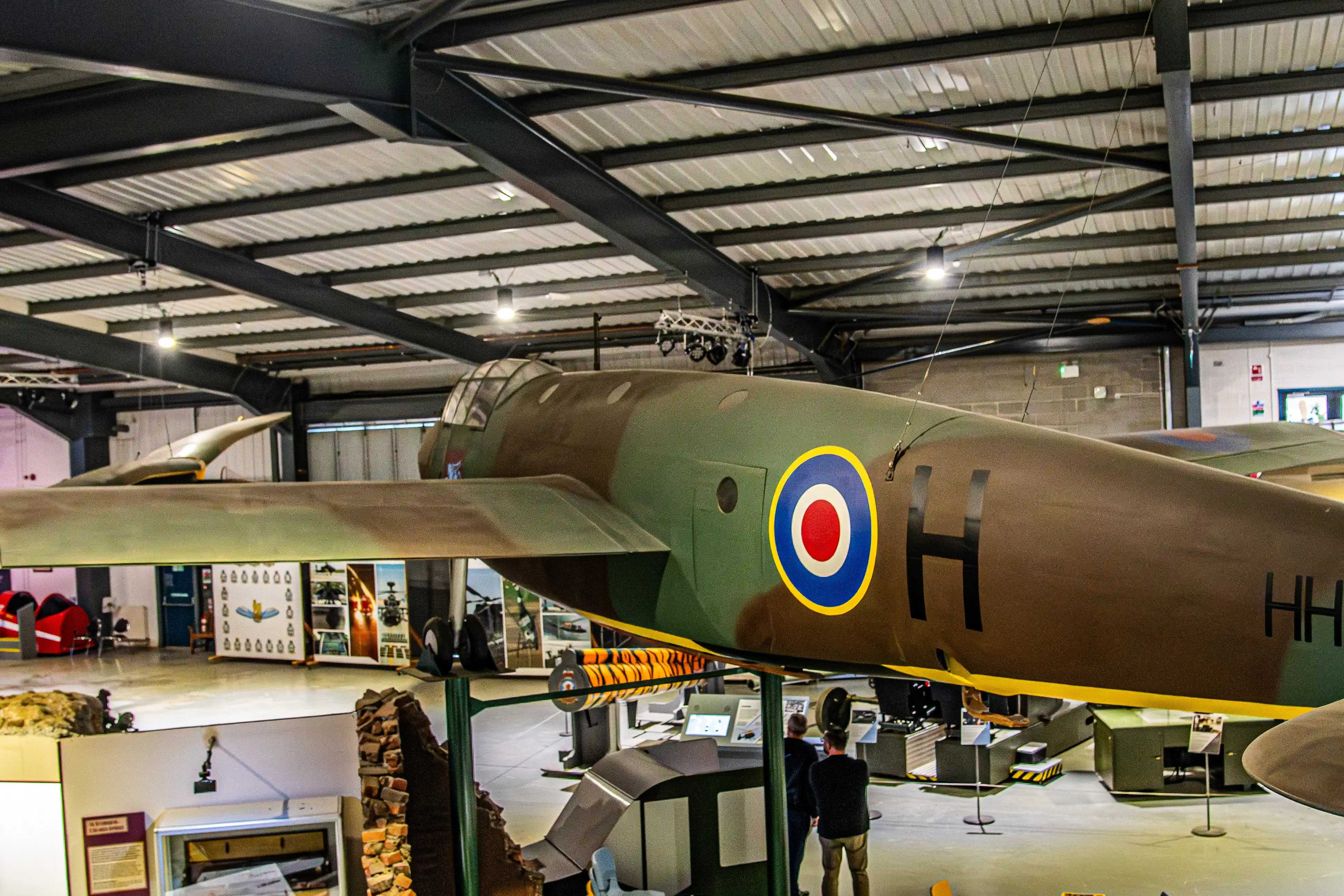 Olive-green aircraft on display in a museum hangar, showing RAF-style roundel and “H” marking along the fuselage.  