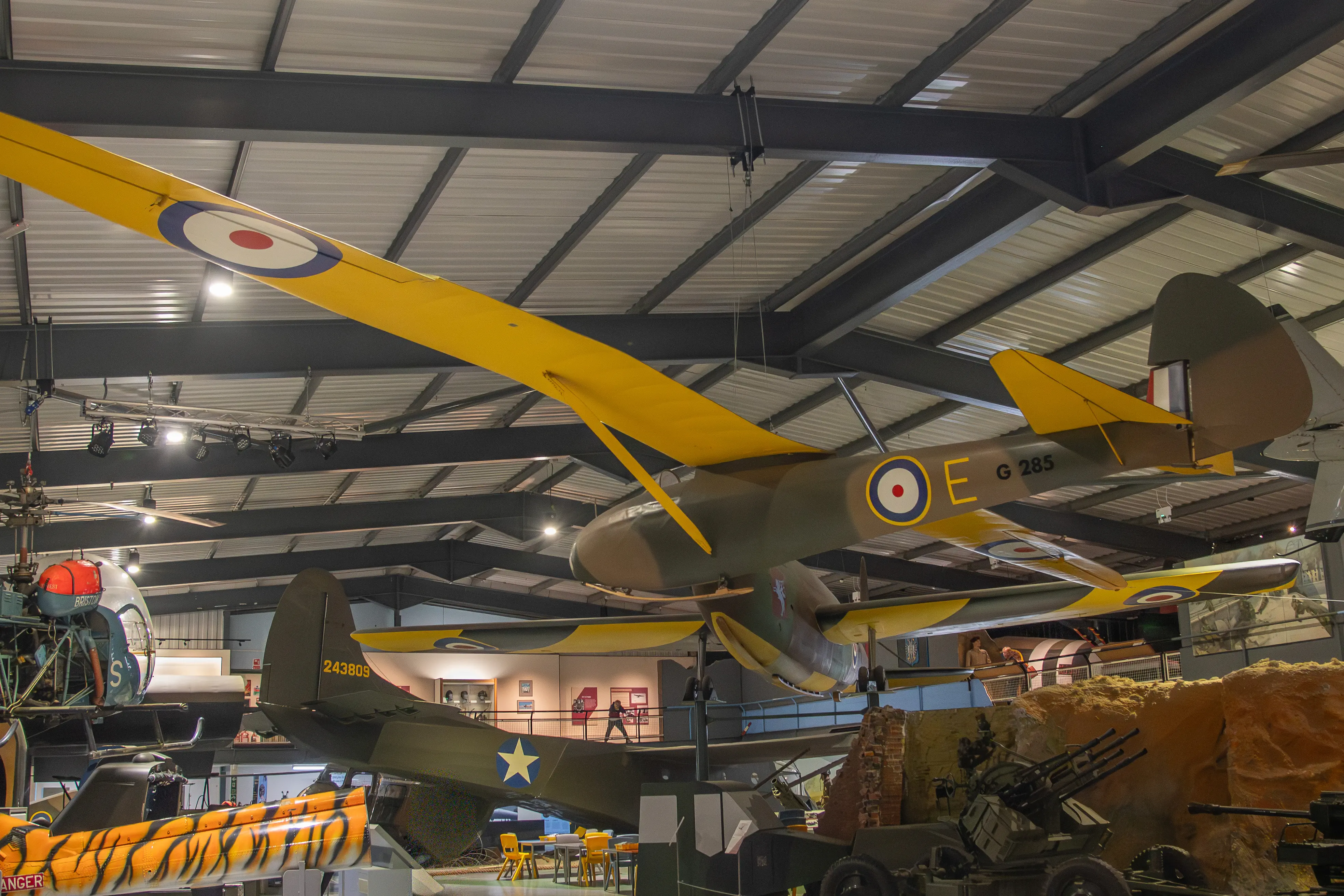 Indoor aviation museum showing a suspended World War II British glider with camouflage and yellow wings, surrounded by other military aircraft and vehicles.