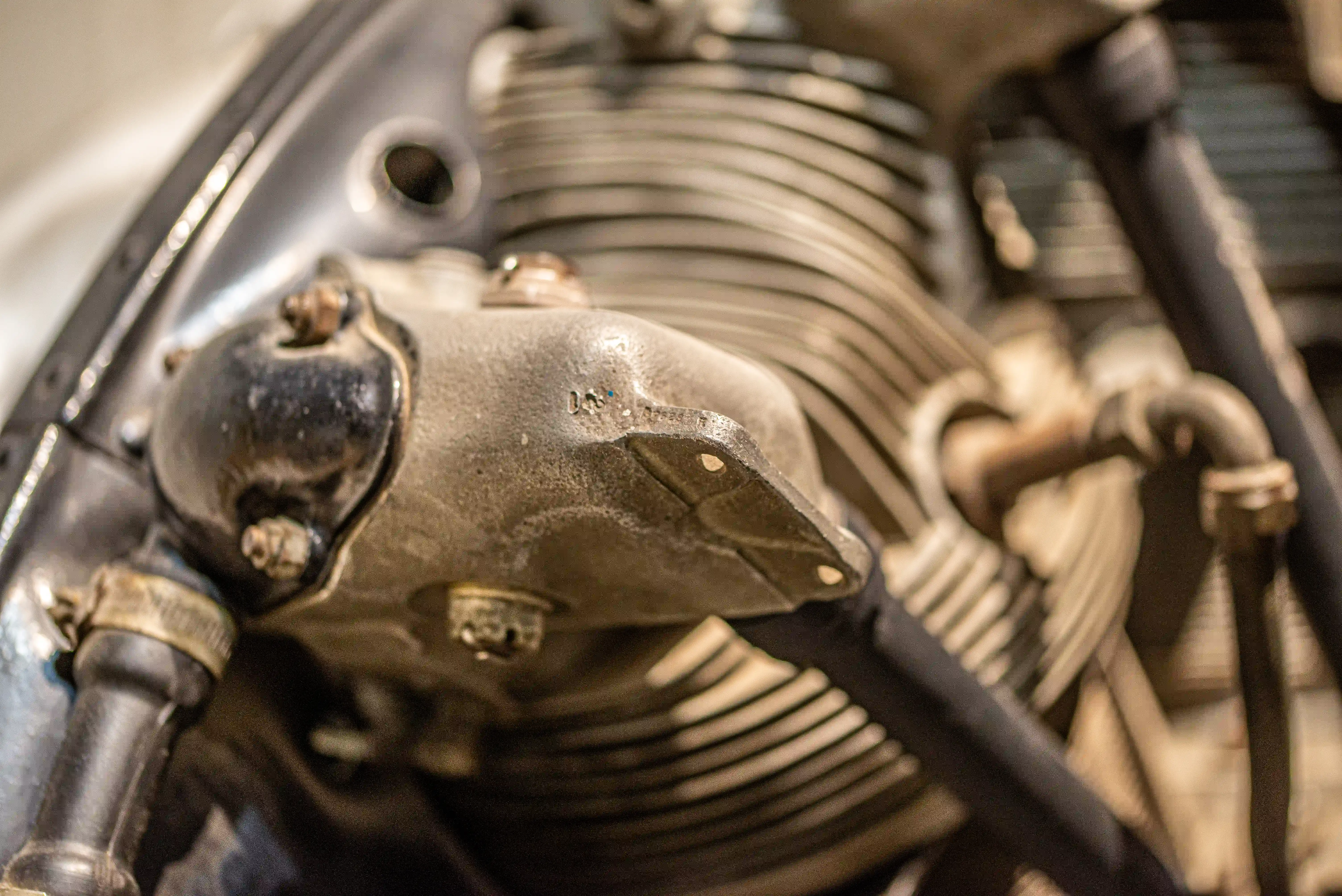 Close-up, slightly blurred image of a vintage aircraft engine's finned cylinder head, showing metal pipes, bolts, and a dark casing.