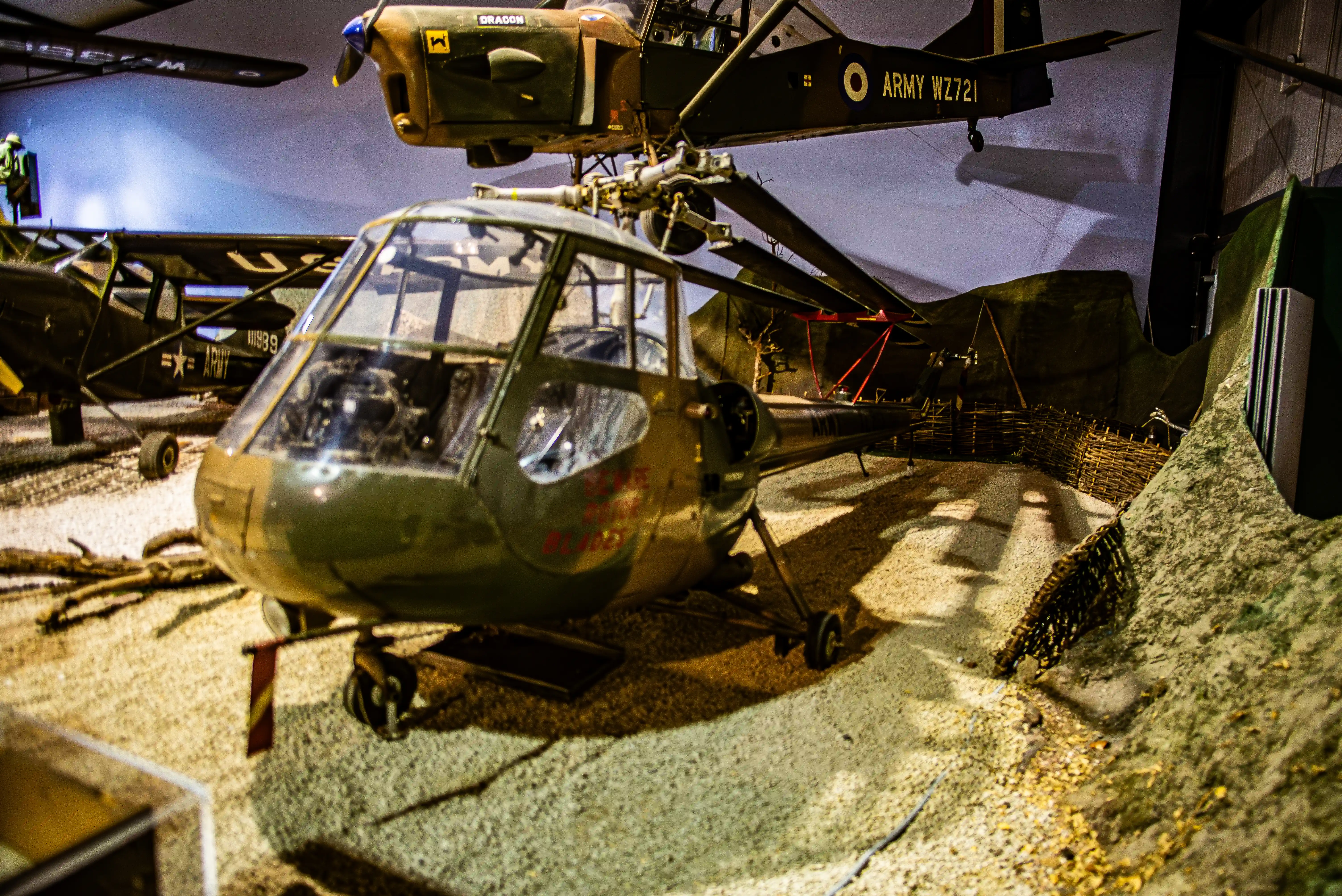 A close-up view of a green military helicopter with a clear cockpit, parked on a gravel-covered floor in a museum exhibit. Other vintage aircraft are visible in the background.