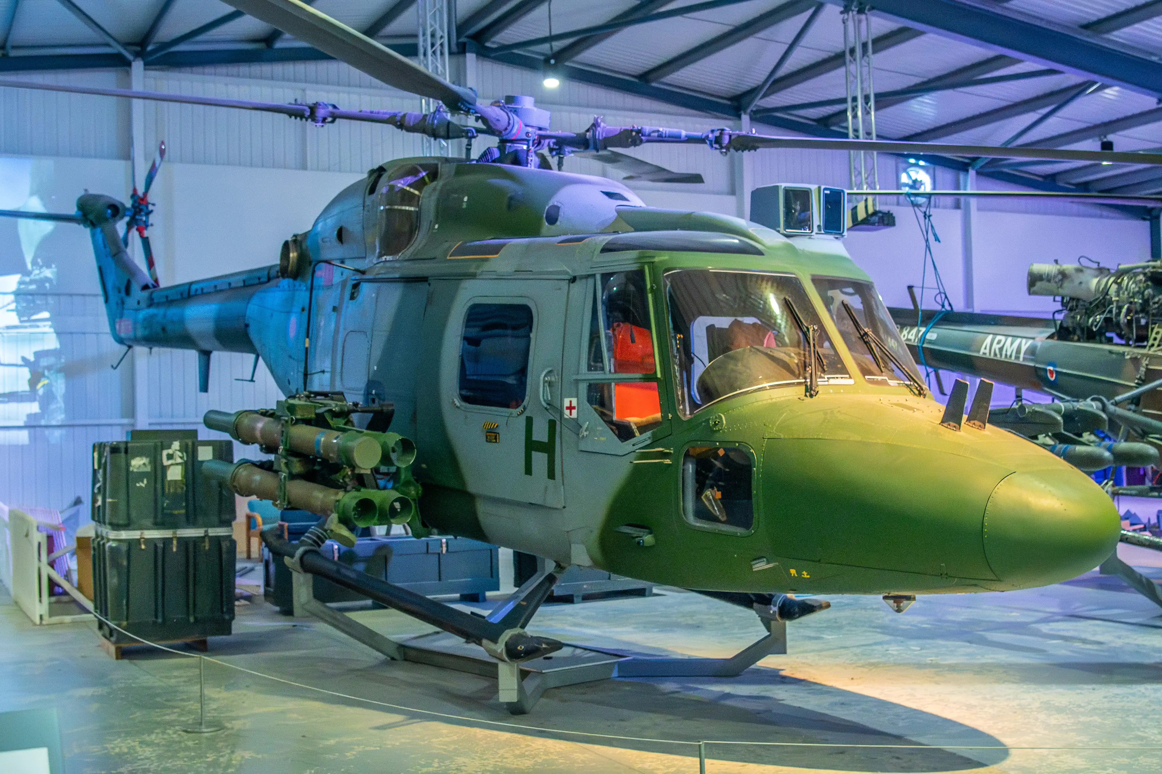 A military helicopter with rotors and missile launchers, painted in camouflage, stands on a display in a hangar.