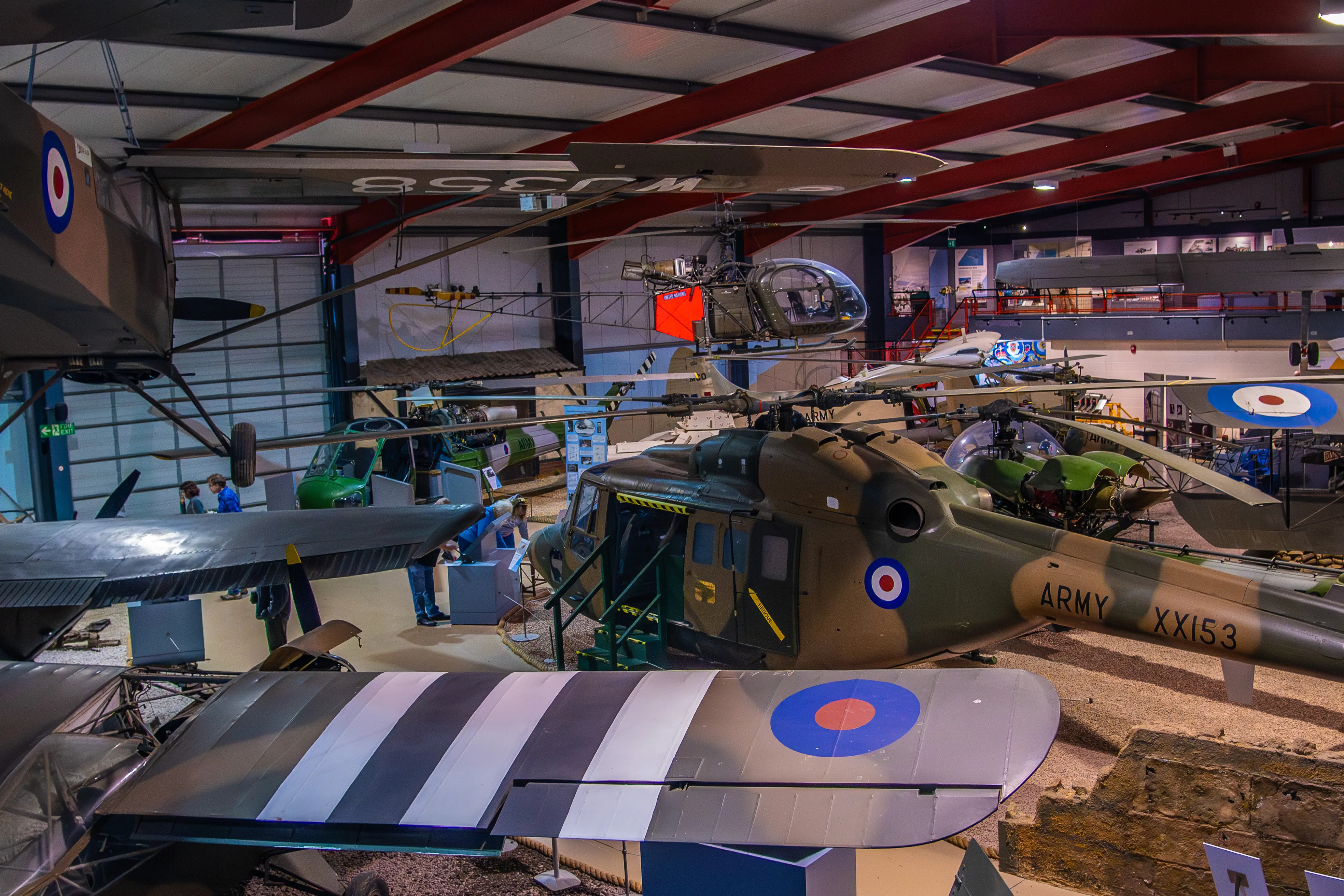 Indoor aviation museum with historic military aircraft and helicopters, including a camouflage helicopter marked “ARMY XX153” and vintage RAF aircraft.