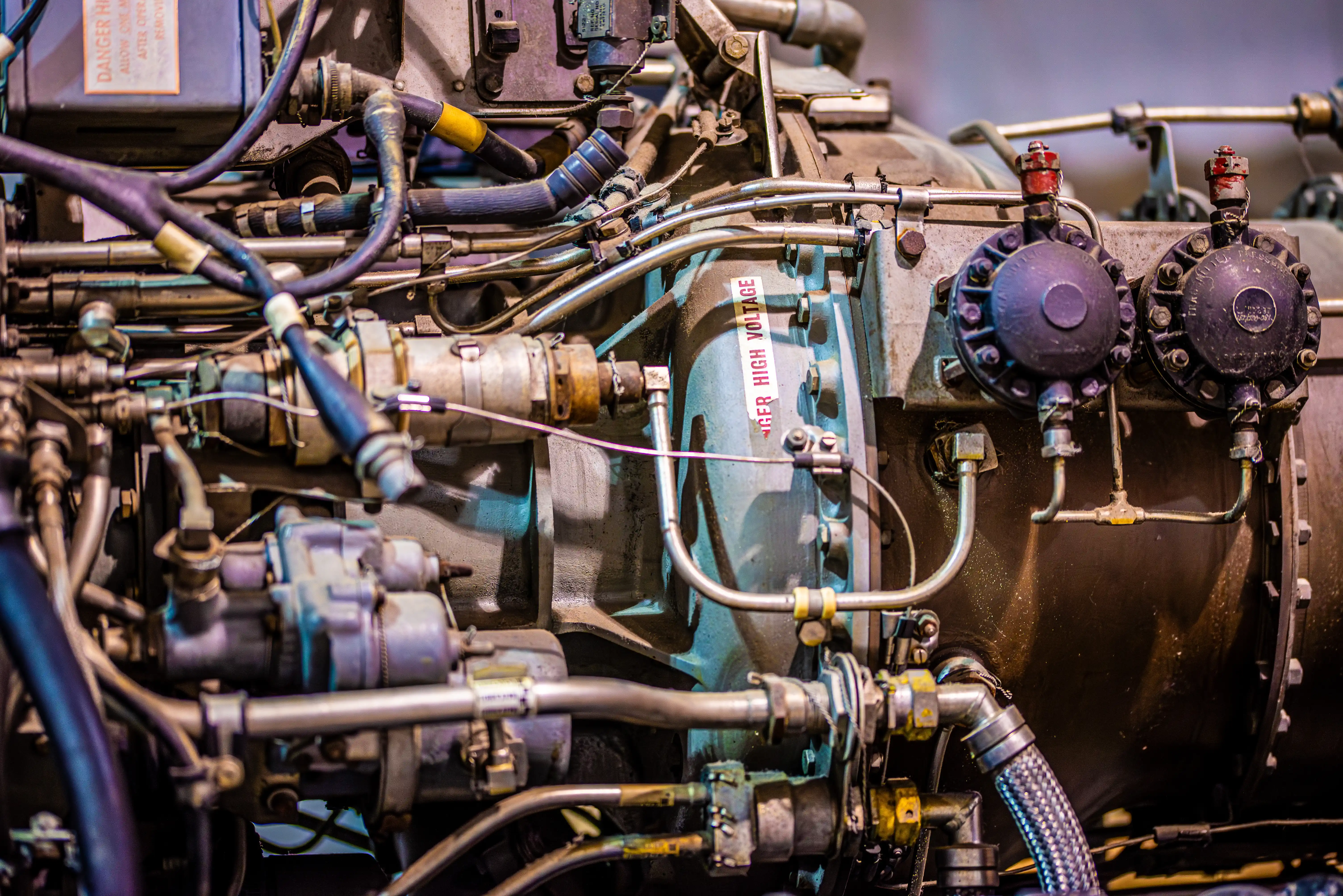 Close-up photograph of a complex engine assembly with metal pipes, valves, electrical cables and a high-voltage warning label.