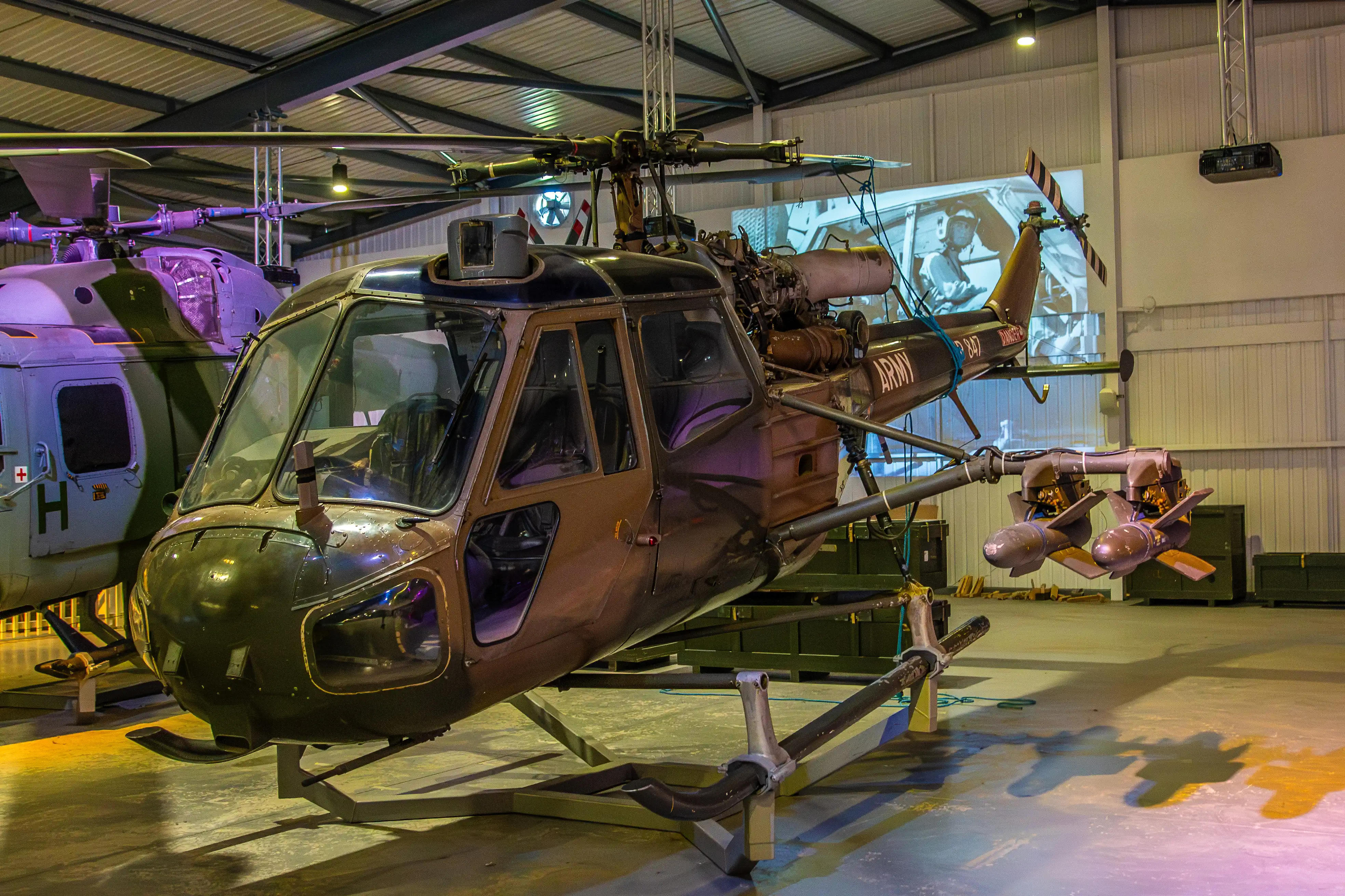 A vintage military helicopter marked with 'ARMY' displayed in a museum hangar. The helicopter has visible rotors, a cockpit, and an exposed engine. The hangar is dimly lit, with other aircraft and exhibits in the background.