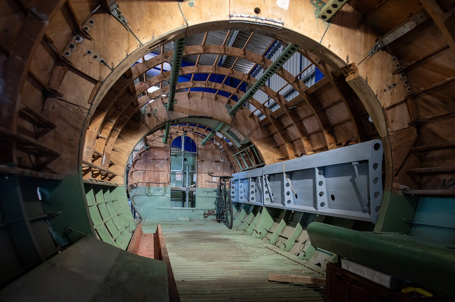 The image depicts the interior of an aircraft fuselage under construction or maintenance. The structure is largely composed of wooden frames and metal components, with visible ribs and support beams. There is a bicycle placed inside the fuselage, likely for scale or transportation purposes. The setting appears to be an industrial or workshop environment, indicated by the unfinished walls and exposed materials.