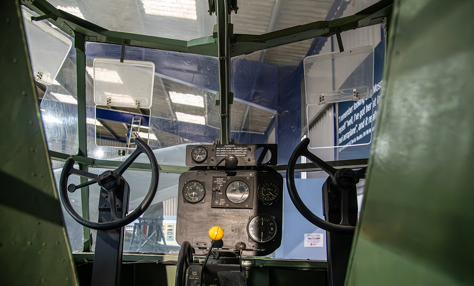 The image shows the interior of an aircraft cockpit. The cockpit features a central control panel with various gauges and dials, a control stick, and a yellow control knob. There are two large windows at the front, providing a view outside. The cockpit appears to be part of a vintage or historical aircraft, possibly on display in a museum.