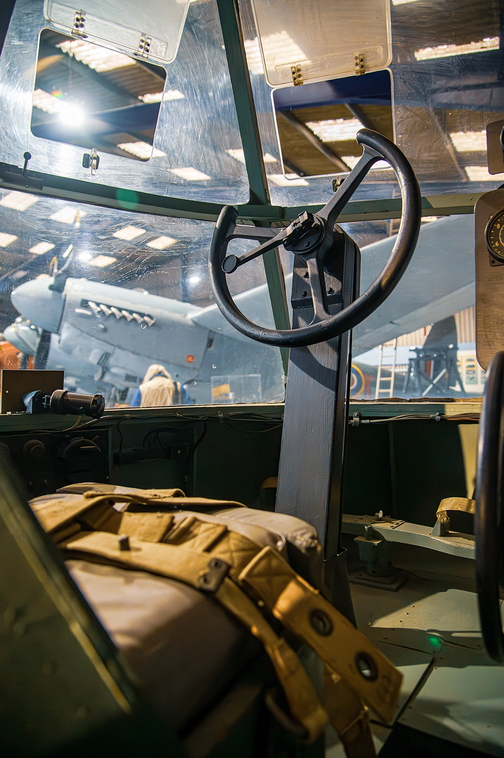 The image depicts the interior of an aircraft cockpit with a clear view of the steering wheel and control panel. The cockpit windows reveal a view of another aircraft outside, suggesting the setting is likely an aviation museum or an airshow. The seating appears to be upholstered in fabric, and there is a tan-colored bag or cushion in the foreground. The lighting suggests it is daytime, with sunlight streaming through the windows.