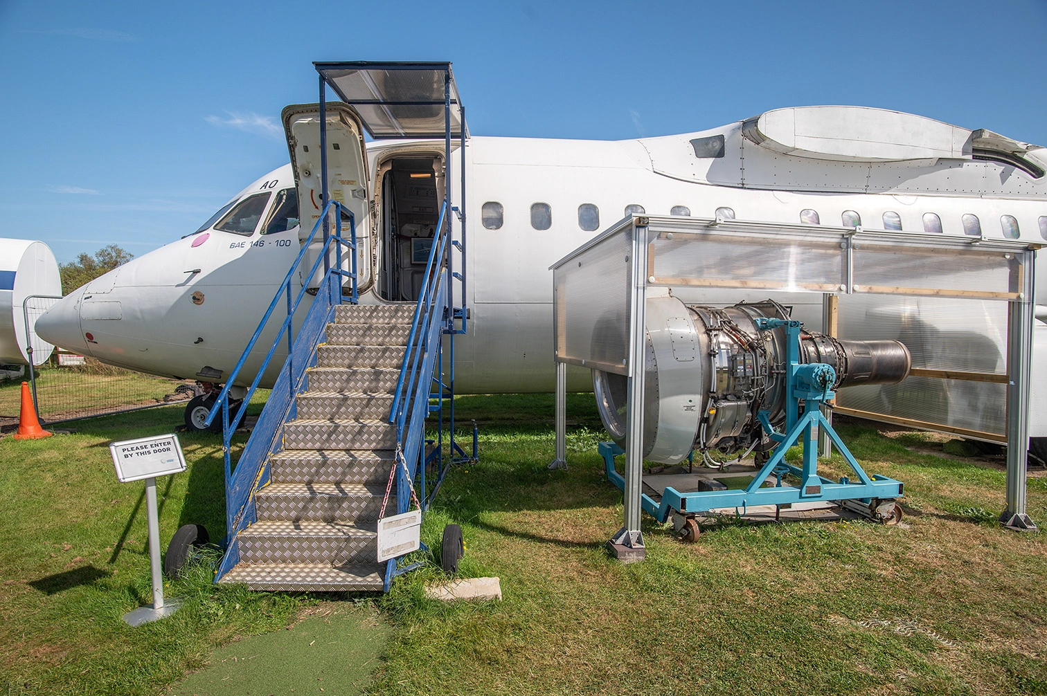 The image depicts an aircraft on display with its entrance staircase extended. The aircraft's door is open, and a sign indicates the entrance point. In front of the aircraft, there is an exposed jet engine mounted on a stand, likely for educational or exhibition purposes. The scene is set outdoors on a grassy area under a clear blue sky.