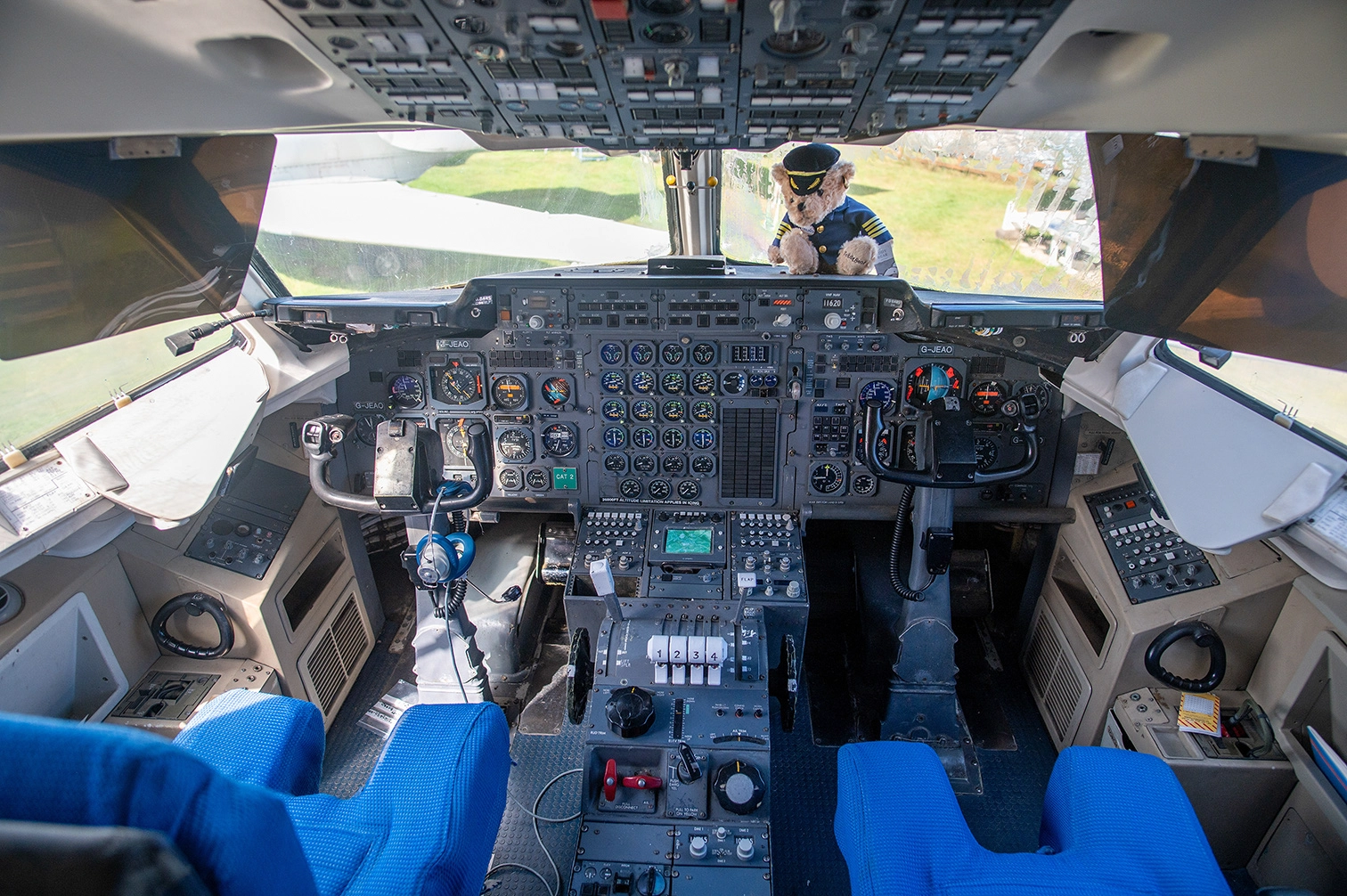 The image depicts the cockpit of an aircraft. It features a complex array of instruments, controls, and displays used for flying the plane. The pilot seats are visible with blue covers, and there is a teddy bear dressed as a pilot placed on the dashboard, visible through the windshield. The cockpit is well-lit, showing a clear view of the runway outside.