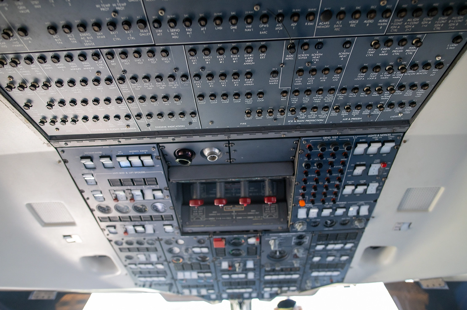 The image depicts the overhead control panel of an aircraft cockpit. This panel contains numerous switches, buttons, and indicators used for managing various aircraft systems, including engine controls, lights, and other essential functions. The layout is complex and organized into different sections for specific controls and monitoring. It is designed to provide pilots with easy access to critical systems during flight.