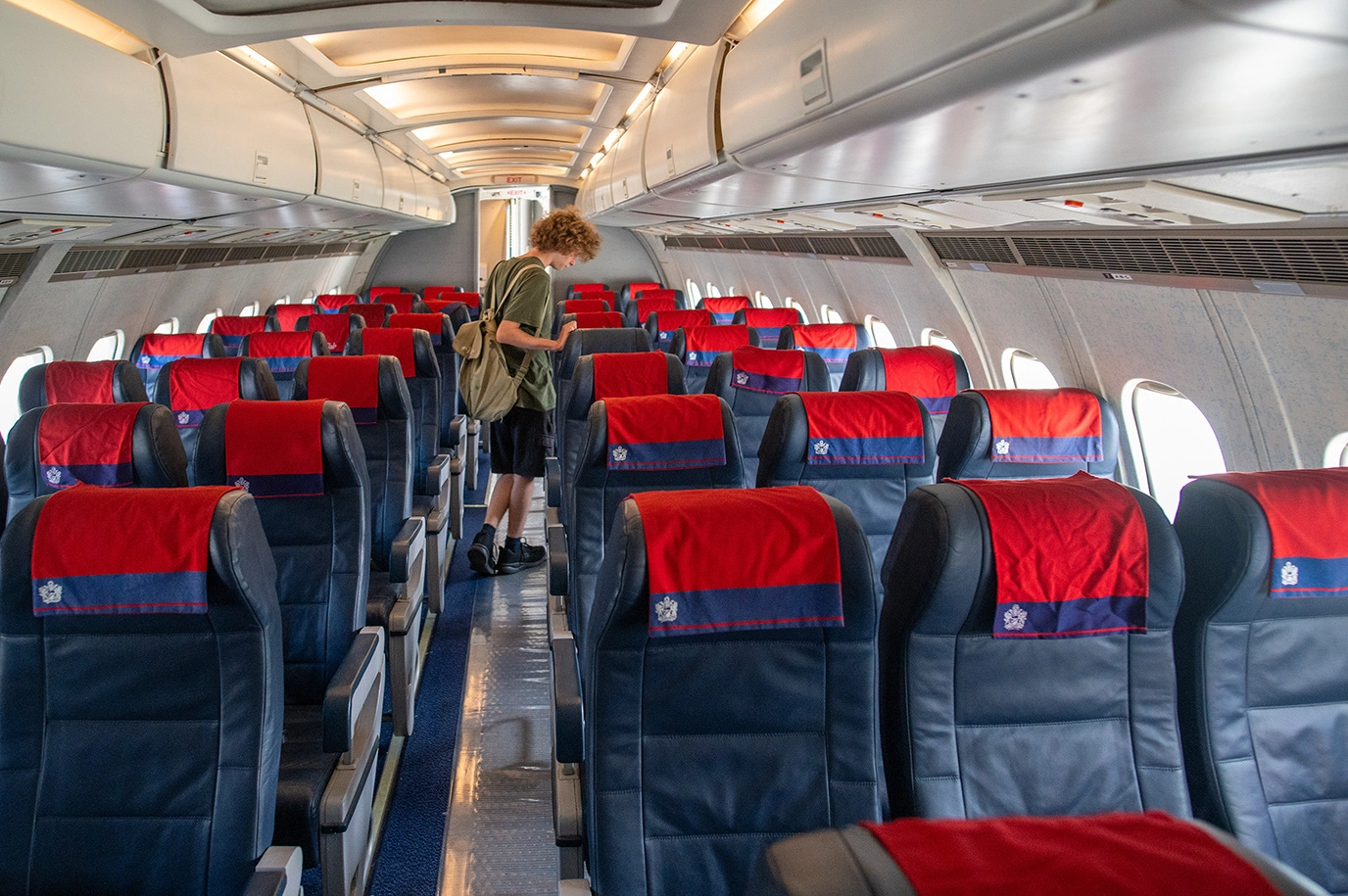 The image depicts the interior of an airplane cabin. The seats are arranged in rows with red and blue upholstery. A person with curly hair is seen walking down the aisle, carrying a backpack and looking at their phone. The cabin appears to be mostly empty, with overhead compartments and windows visible along the sides.