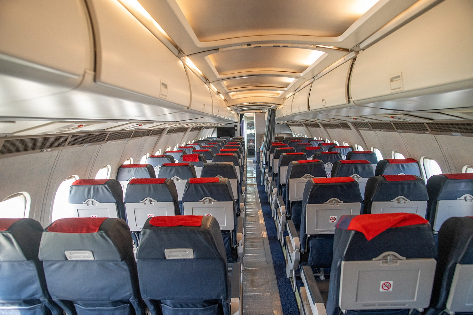 The image depicts the interior of an airplane cabin. The seats are arranged in a typical configuration with a central aisle. Each seat has a red headrest cover and a tray table at the back. The overhead compartments are visible, and the cabin appears to be empty with no passengers present.