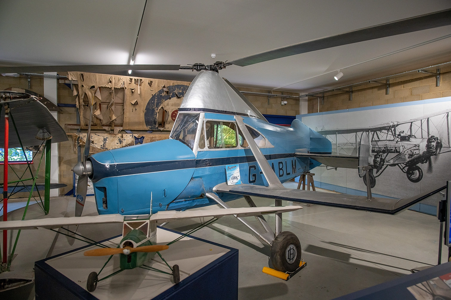 The image depicts an indoor aviation museum exhibit featuring several historical aircraft. The central focus is a light blue aircraft with the registration G-ABLM. In the background, there is a black and white photograph of a vintage biplane. The exhibit includes various other aircraft and aviation-related displays, creating an educational and visually engaging environment.