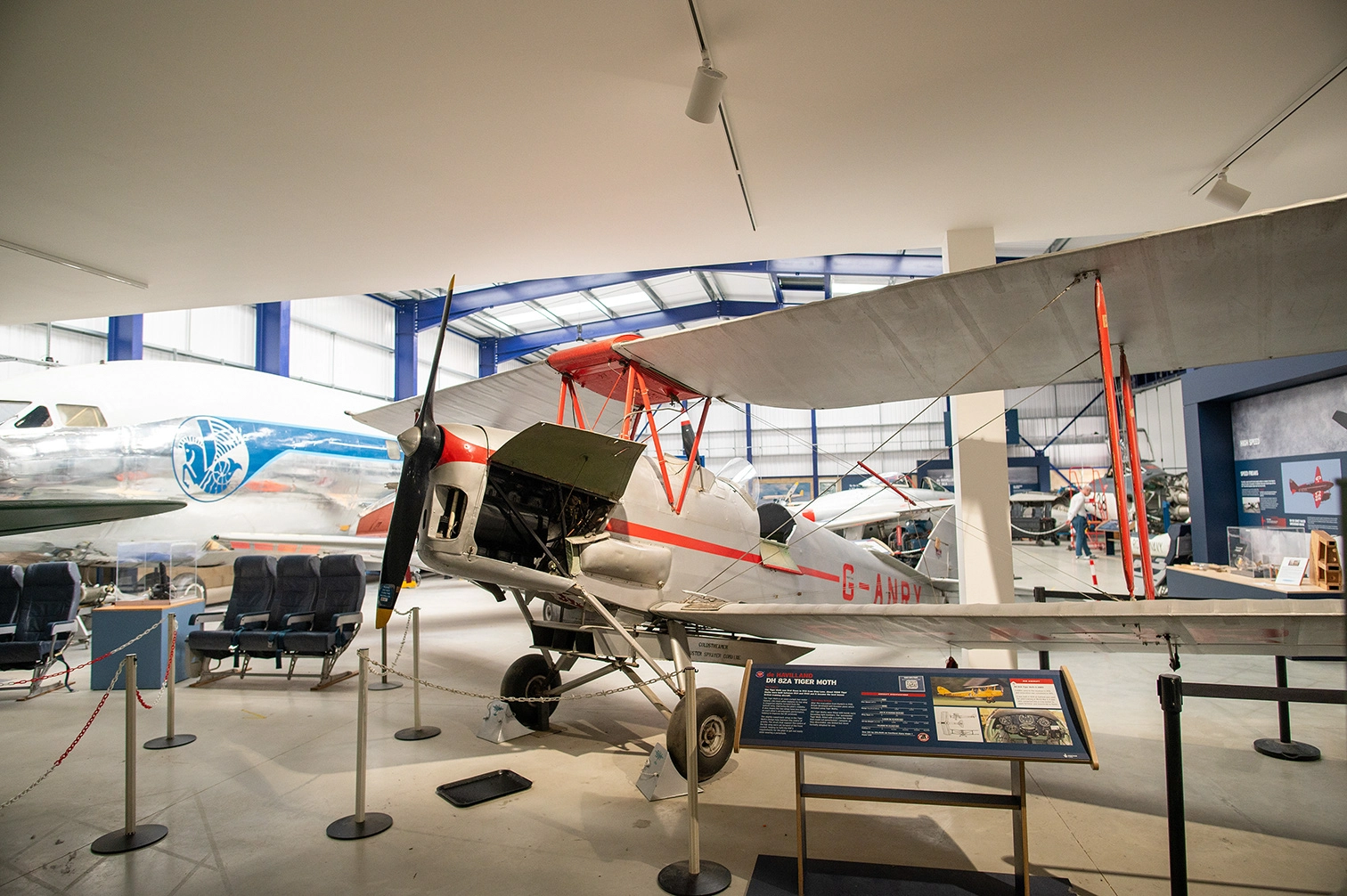 The image depicts an indoor aviation museum exhibit. The focal point is a vintage biplane, specifically a DH 82A Tiger Moth, displayed prominently in the center. Surrounding the aircraft are informational displays and seating arrangements, likely for educational purposes. Other aircraft and exhibits can be seen in the background, indicating a comprehensive collection of aviation history.