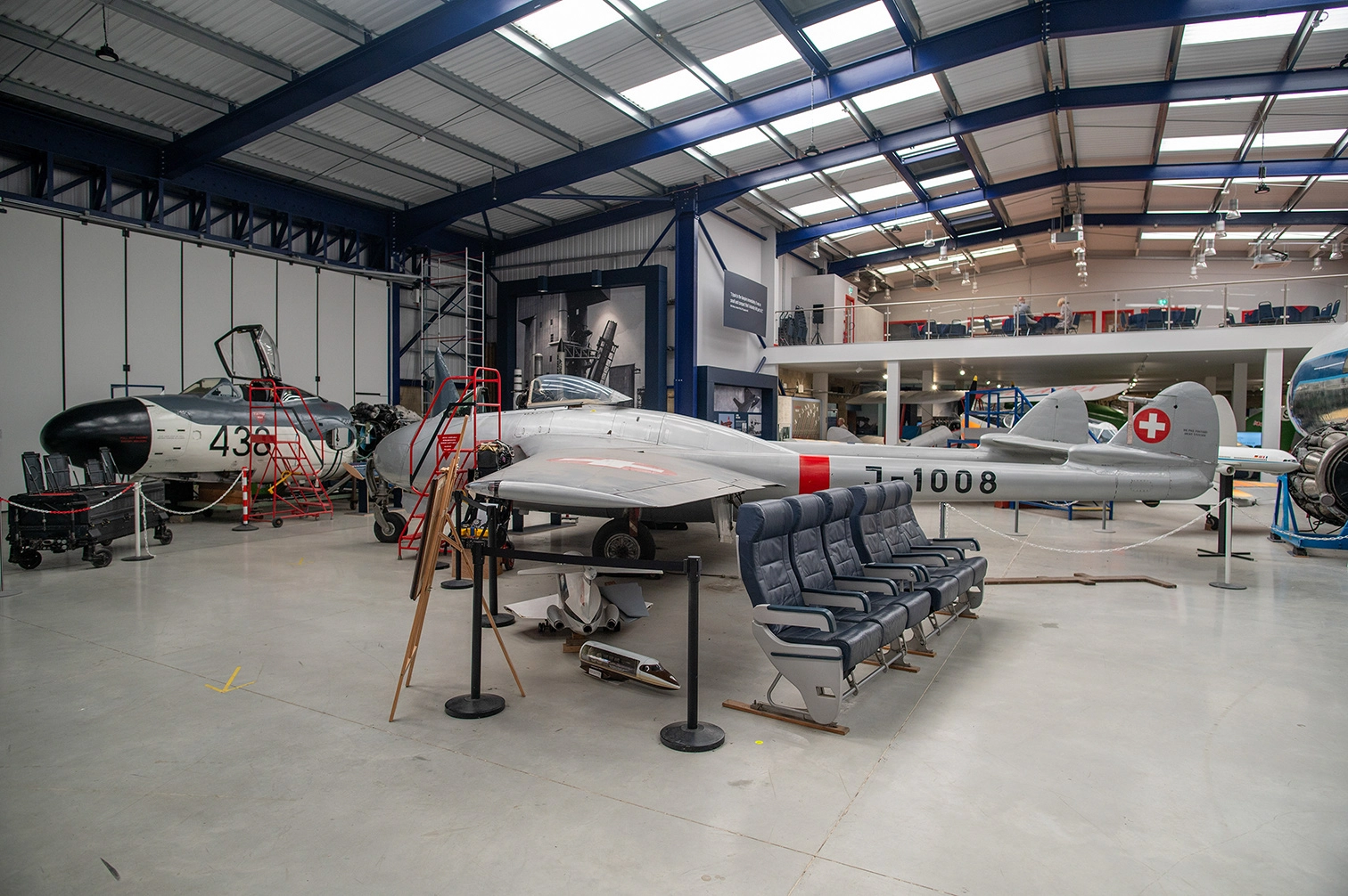 The image depicts an indoor aviation museum or hangar. Several aircraft are displayed, including a prominent silver jet with a red cross and the number J-1008. There are also seating arrangements and various equipment on display. The area is well-lit with a high, industrial-style ceiling.