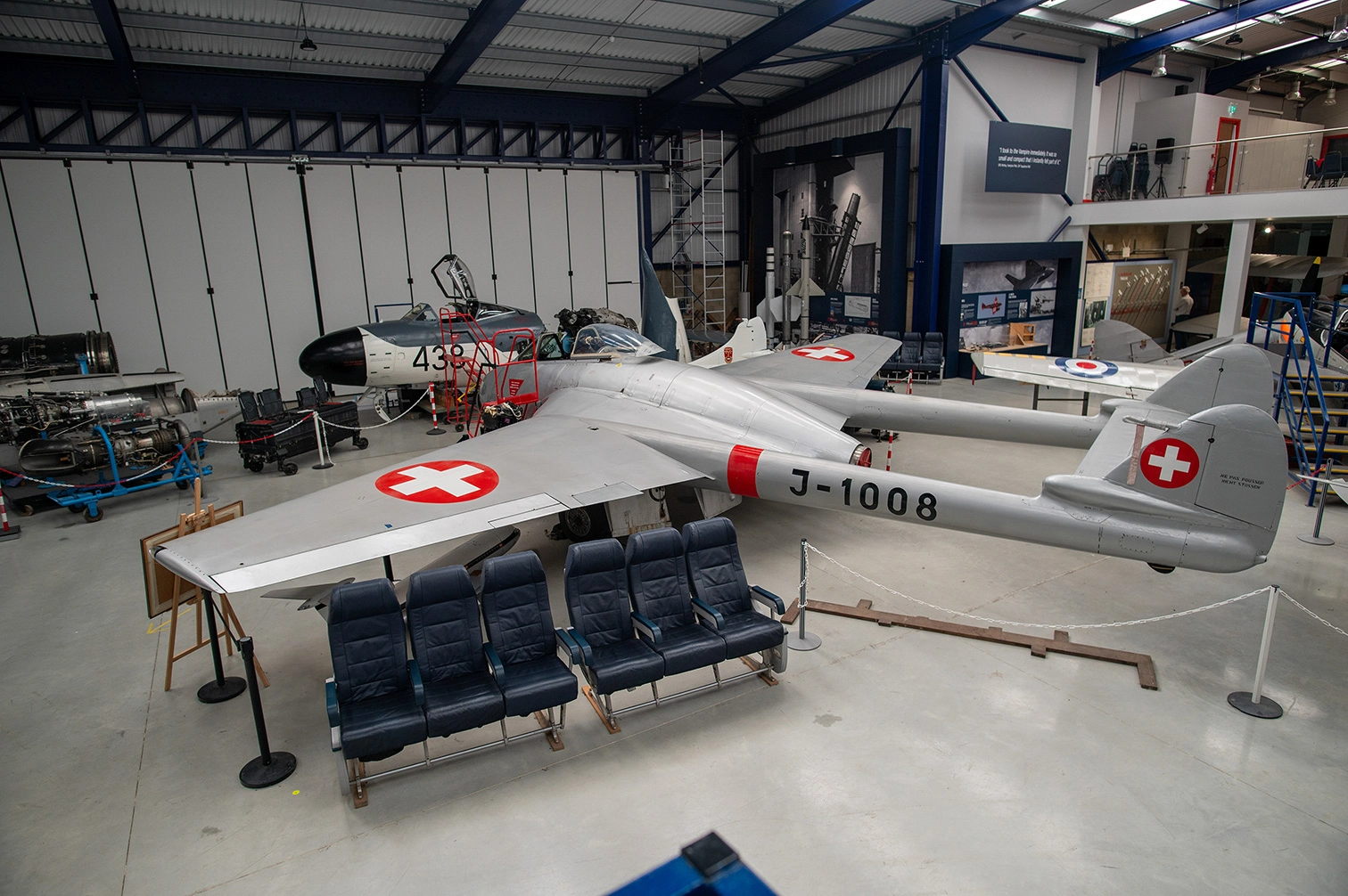 The image depicts an indoor aviation museum or hangar. The central focus is a vintage military aircraft with Swiss markings and the identification number J-1008. The aircraft is surrounded by various other planes and aviation equipment, including engines and other aircraft parts. There are also seating arrangements in front of the main aircraft, likely for educational or tour purposes.