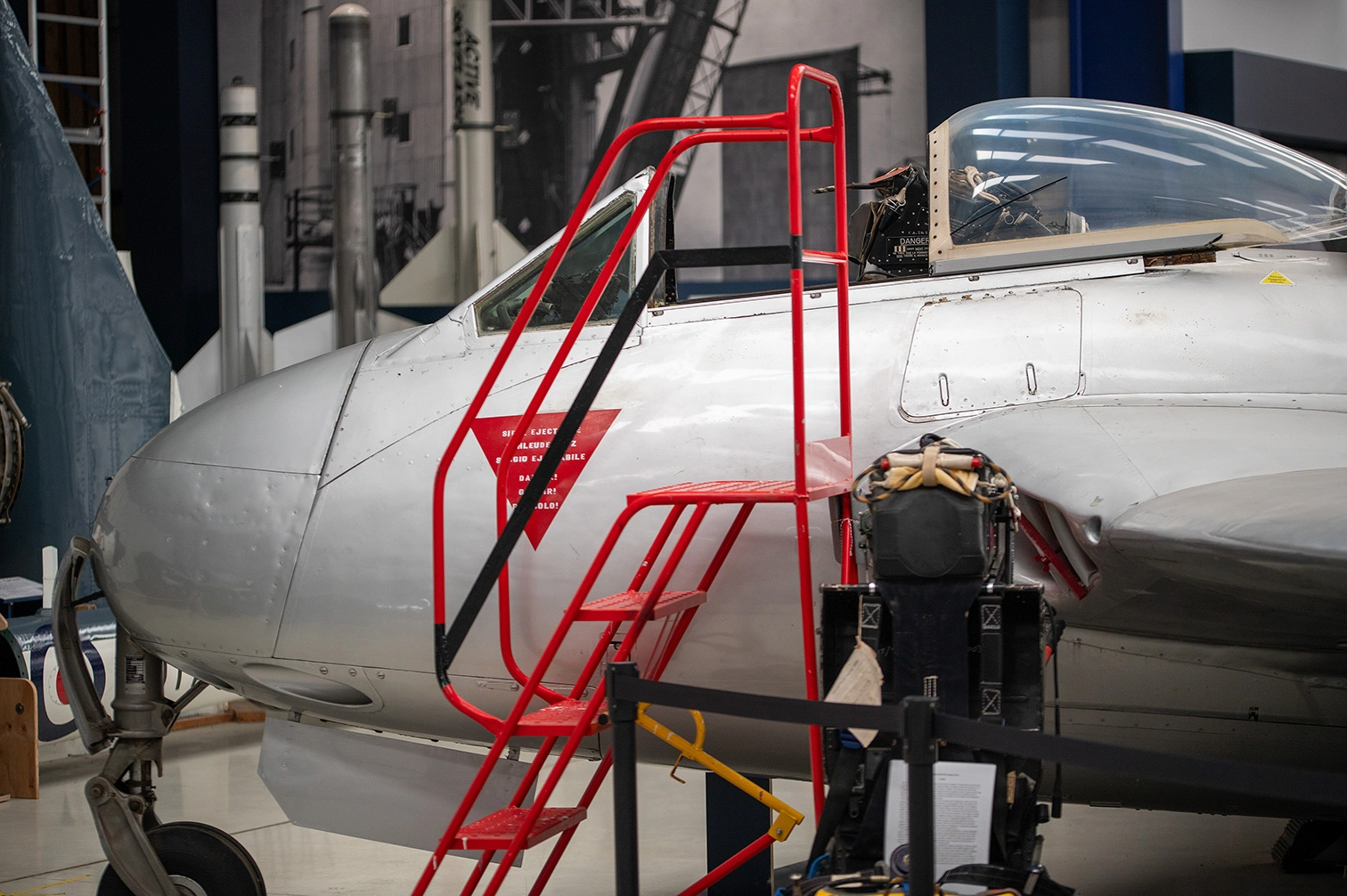 The image depicts a close-up view of a military aircraft inside a hangar or museum. The aircraft features a cockpit with a transparent canopy, and a red ladder is positioned next to it, likely for access. The aircraft's nose has various markings and text, and there is an ejection seat visible through the canopy. The surrounding area includes other aircraft and industrial equipment, indicating a display or maintenance setting.