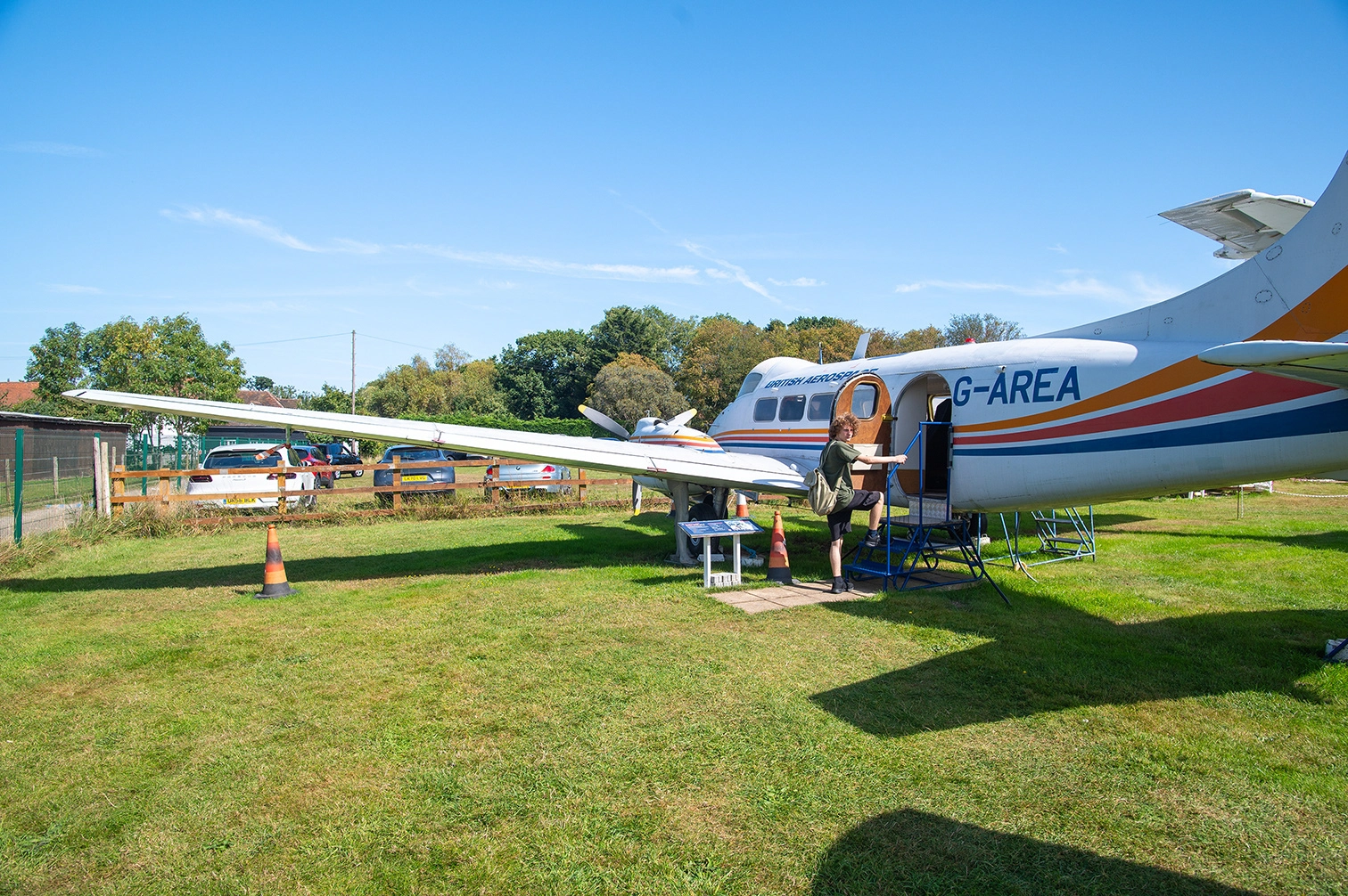 The image shows a small aircraft on display outdoors, with a person standing on a ladder near the open door. The aircraft is painted with colorful stripes and has the text 'G-AREA' visible on its side. The scene appears to be part of an aviation exhibit or airshow, with several cars parked in the background and a grassy field in the foreground. The weather is clear and sunny.