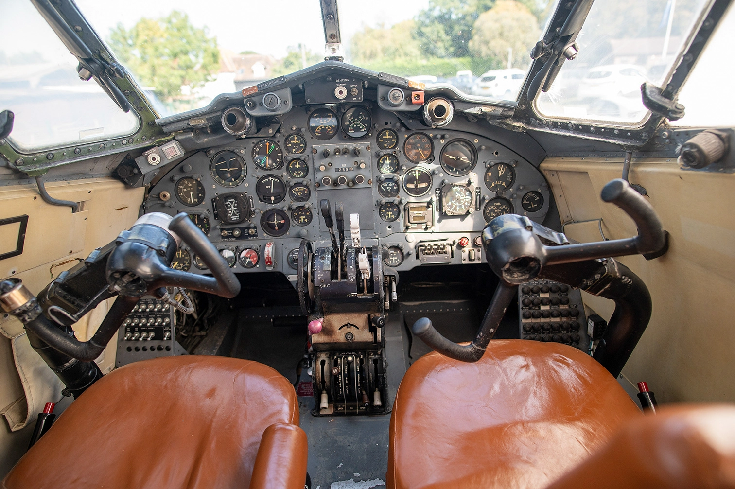 The image depicts the interior of an aircraft cockpit. It features a complex array of instruments and controls, including various gauges, dials, and switches on the dashboard. There are two leather seats, one of which is prominently visible in the foreground. The cockpit has a vintage appearance, suggesting it might be from an older aircraft model. The view outside shows a bright day with some greenery and vehicles visible through the windshield.