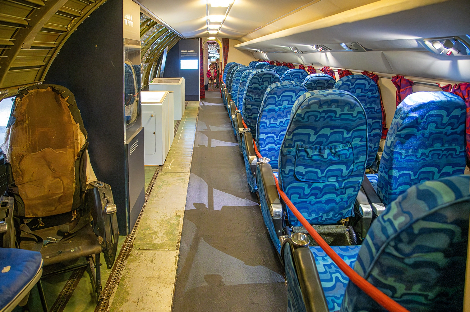 The image depicts the interior of an airplane cabin. The seats are arranged in rows, with a mix of worn-out and well-maintained upholstery. The left side shows a damaged, brown seat, while the right side features blue patterned seats in better condition. The cabin includes overhead compartments and a narrow aisle leading towards the back of the plane.