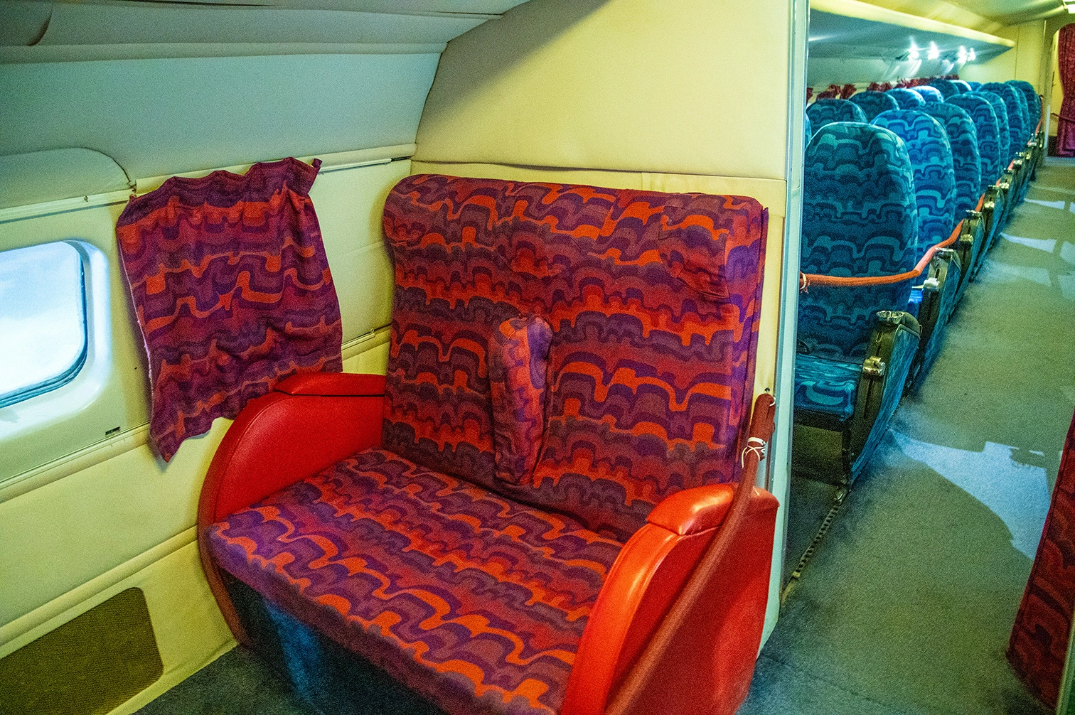 The image depicts the interior of an airplane cabin. The seats in the foreground are upholstered with a vibrant, patterned fabric in shades of red and purple. A red blanket is draped over one of the seats, and the seats themselves have red armrests. In the background, rows of blue seats are visible, arranged in a typical airplane seating configuration.