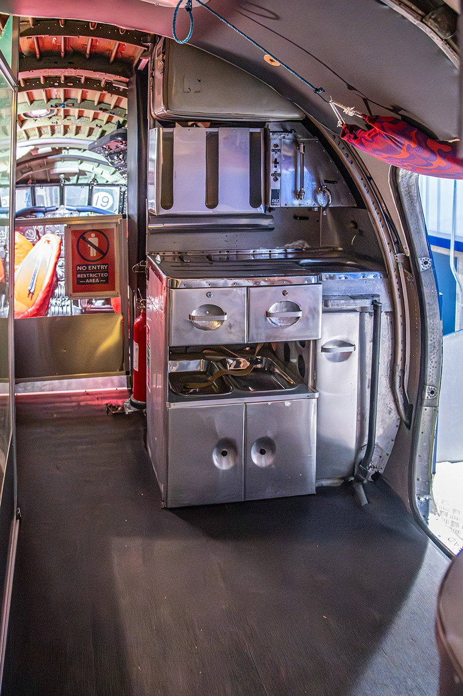 The image depicts the interior of an aircraft, specifically focusing on the galley area. It features metallic storage compartments and kitchen equipment, including an oven. There is a fire extinguisher visible, and a sign indicating a restricted area. The space appears well-organized and designed for food preparation and storage during flights.