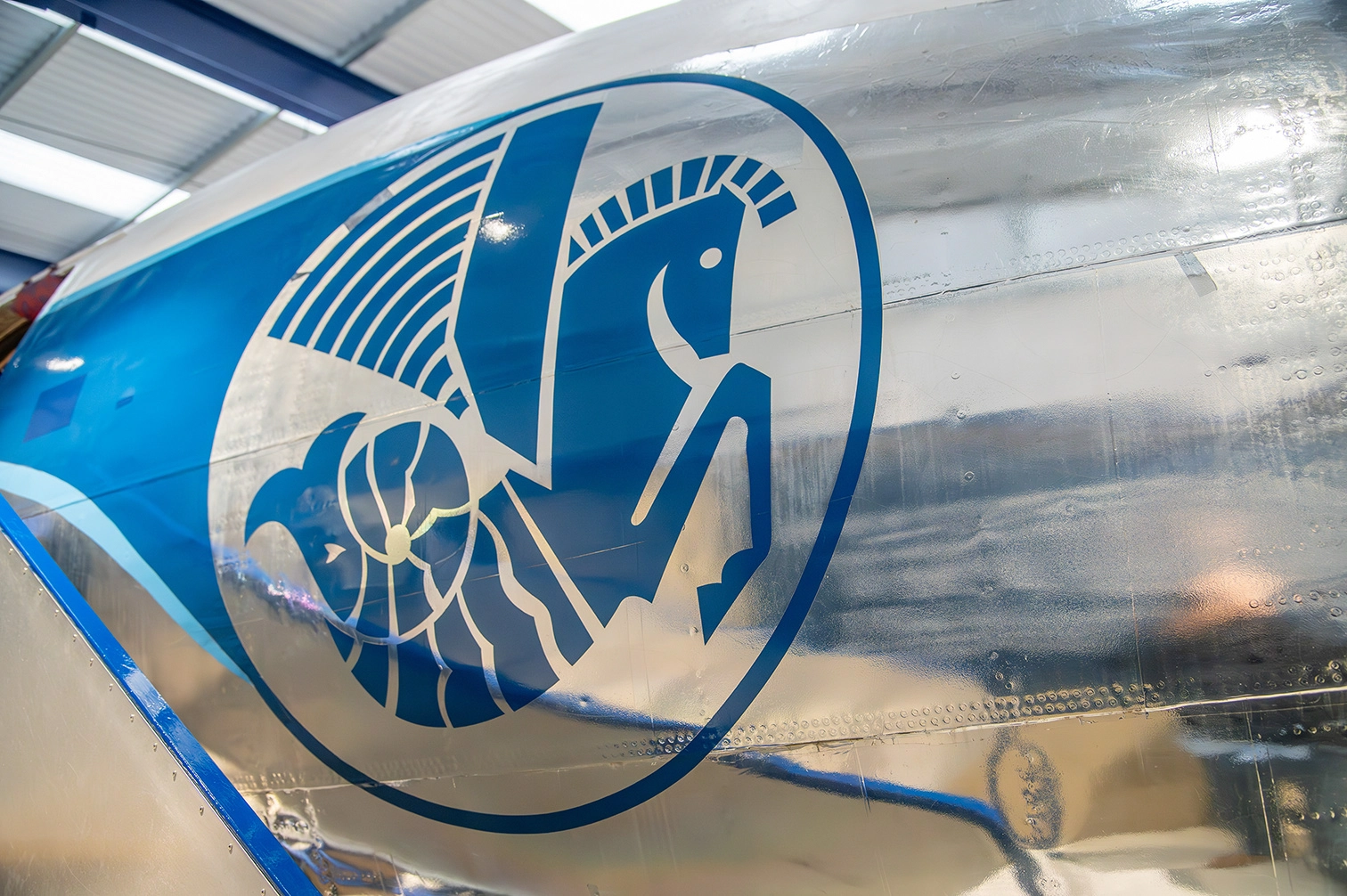 The image shows a close-up view of an aircraft's metallic body, featuring a distinctive blue and white emblem. The emblem includes a stylized design with elements resembling a horse's head, a wheel, and abstract shapes. The aircraft's polished metal surface reflects light, adding a sense of depth to the image. The setting appears to be indoors, possibly in a hangar or museum
