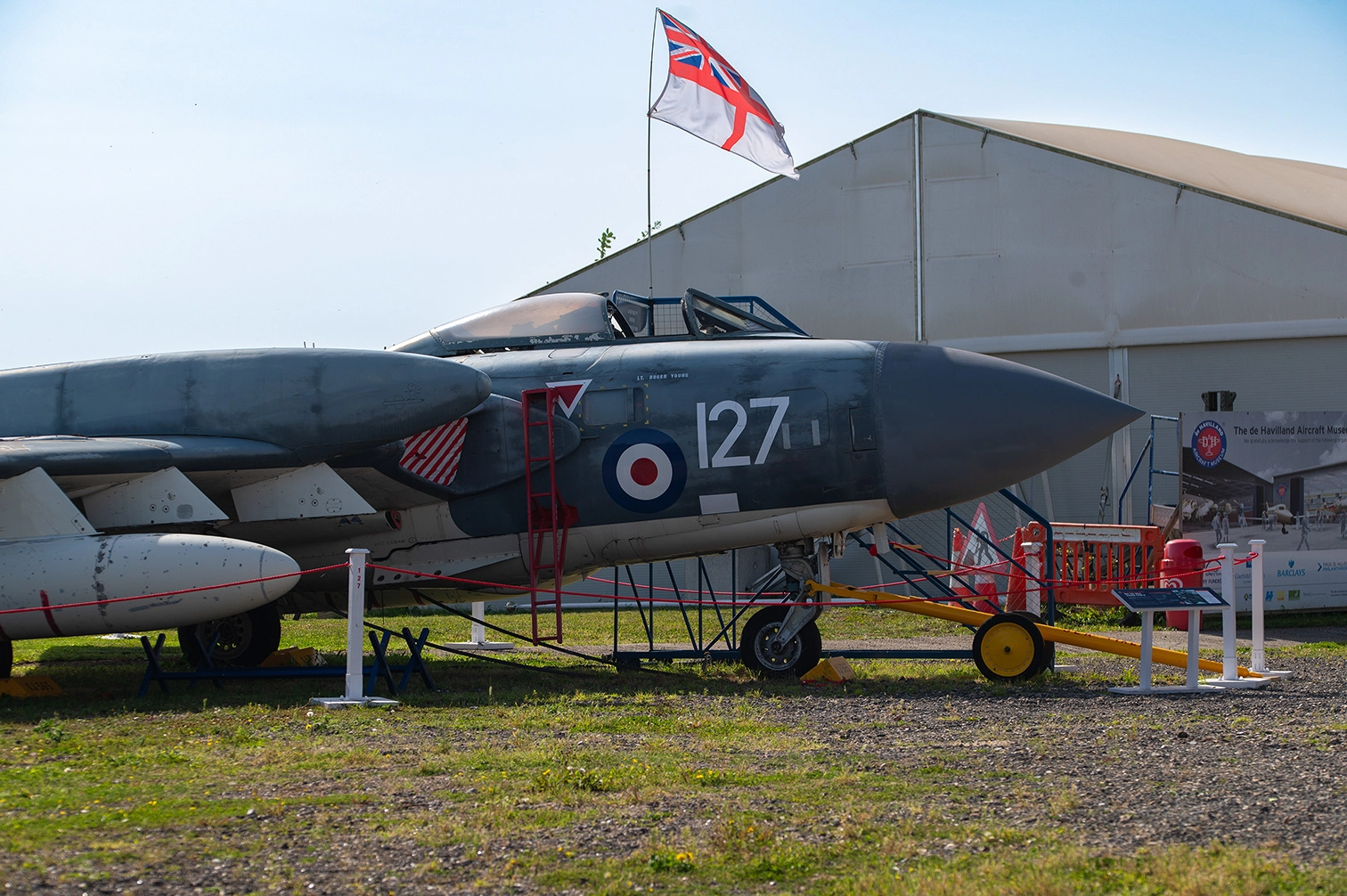 The image depicts an outdoor scene featuring a vintage military aircraft on display. The aircraft is mounted on a platform with stairs leading to its cockpit, and a British flag is flying nearby. Two cars are parked in front of the aircraft, and there are a few people in the vicinity. The setting appears to be a museum or an airshow, with a clear blue sky and greenery in the background.