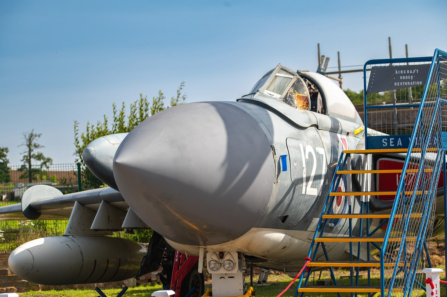 The image depicts a military aircraft undergoing restoration. The aircraft is marked with the number 127 and is surrounded by a protective fence and staircase for access. It is situated outdoors, with visible greenery and a clear sky in the background. The sign nearby indicates that the aircraft is under restoration.