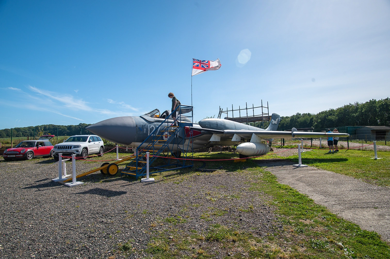 The image depicts an outdoor scene featuring a vintage military aircraft on display. The aircraft is mounted on a platform with stairs leading to its cockpit, and a British flag is flying nearby. Two cars are parked in front of the aircraft, and there are a few people in the vicinity. The setting appears to be a museum or an airshow, with a clear blue sky and greenery in the background.