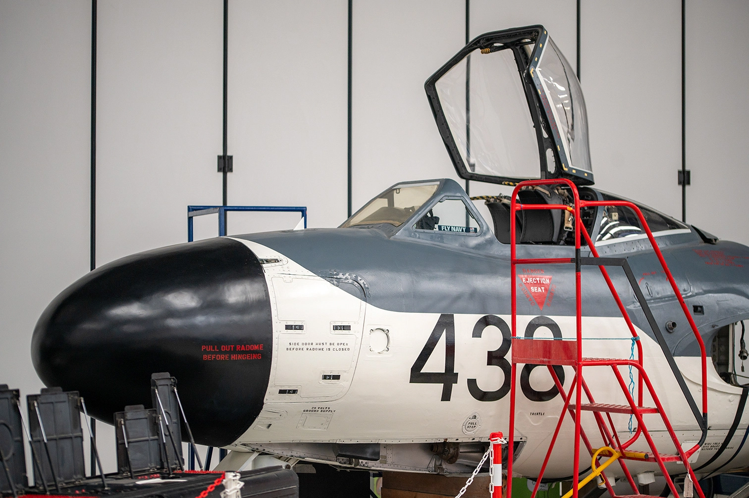 The image depicts a military aircraft inside a hangar. The aircraft has the number 438 painted on its side and features a prominent nose cone. There is a red ladder positioned next to the open cockpit, and various instructions and warnings are visible on the aircraft's body. The aircraft appears to be a vintage or historical model, likely used by the Navy.