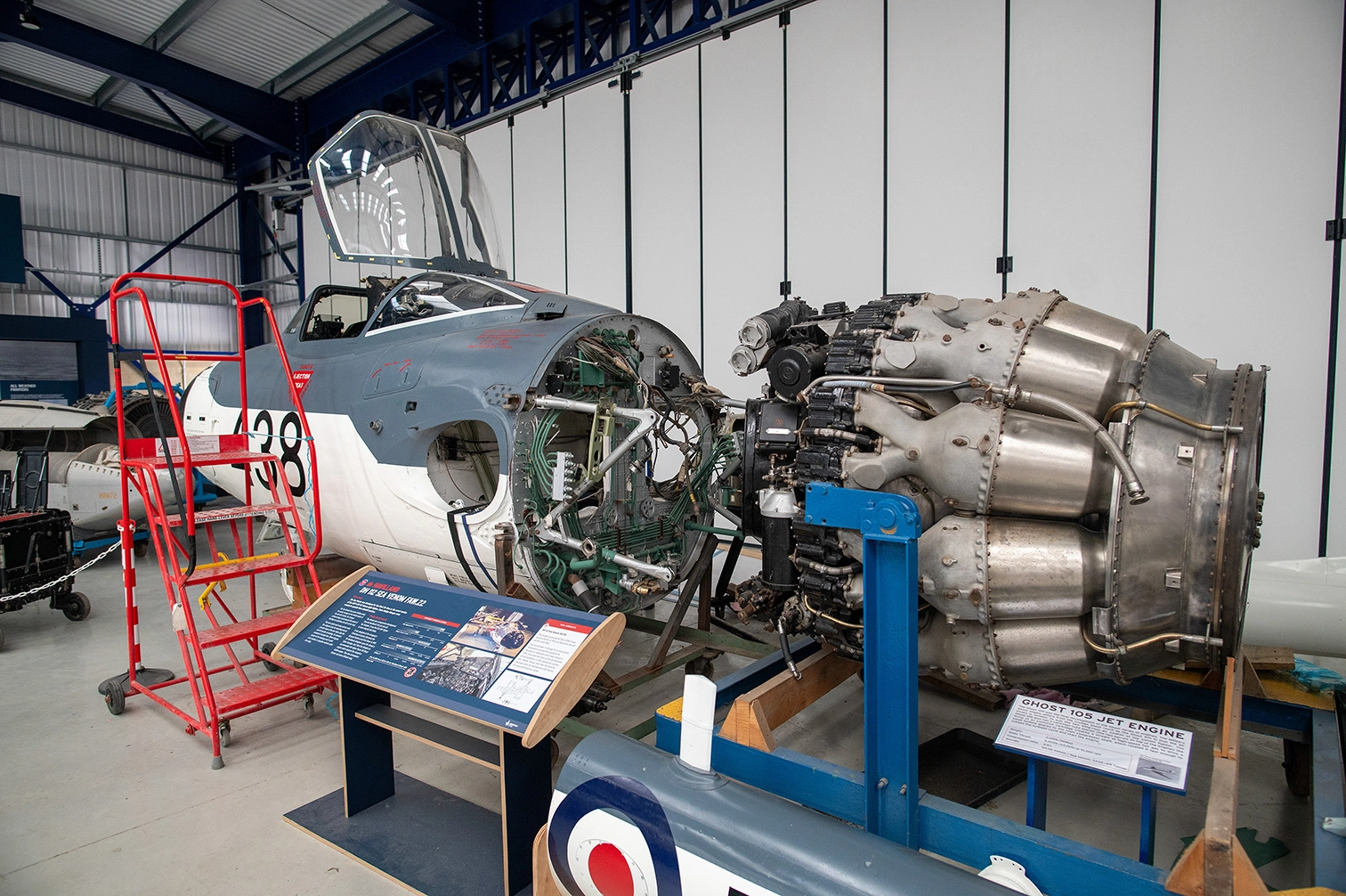 The image depicts an aircraft exhibit in a museum or hangar. A jet aircraft with its engine exposed is prominently displayed, accompanied by informational plaques. The aircraft is marked with the number 38 and has a visible cockpit and intricate engine components. A red ladder is positioned next to the aircraft, and another aircraft is visible in the background. The exhibit appears to be educational, focusing on the engineering and history of the aircraft.