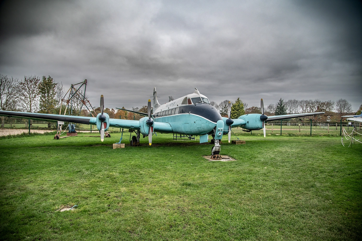 The image depicts an old, stationary airplane displayed on a grassy field. The aircraft appears to be a vintage model with two propellers, and it is situated in an open area with a cloudy sky overhead. The plane is surrounded by a fence and there is some equipment or structures visible in the background. The scene suggests it might be part of an outdoor museum or a display area for historical aircraft.