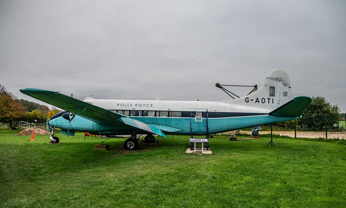 The image depicts a vintage Rolls-Royce airplane with the registration G-AOTI, displayed on a grassy field. The aircraft is painted in a turquoise and white color scheme and appears to be part of a static exhibition. The setting is outdoors with a cloudy sky and some trees in the background.