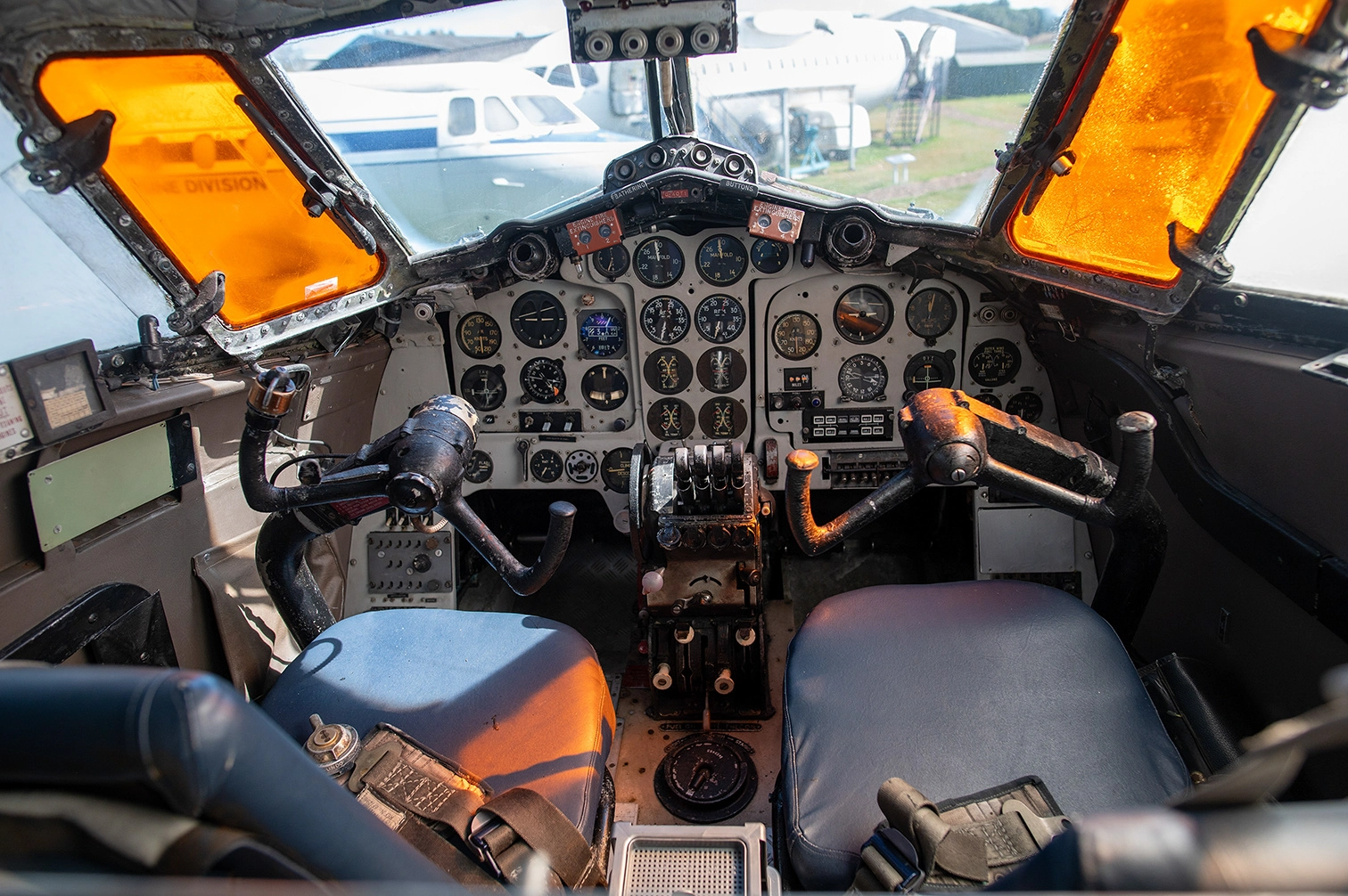 The image depicts the interior of an aircraft cockpit, featuring a complex array of instruments and controls. The cockpit has two seats, one of which is visible, and is equipped with numerous gauges, dials, and switches on the dashboard. The windows are framed with orange covers, and the control yokes are centrally positioned. The overall setting suggests it is an older model aircraft, possibly used for training or historical display.