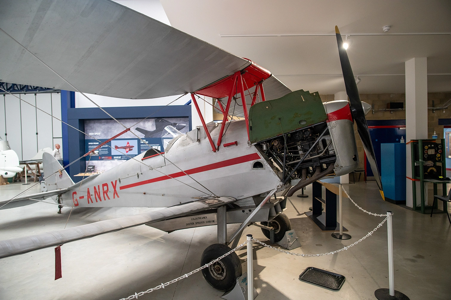 The image depicts a vintage biplane displayed in an aviation museum. The aircraft is suspended on supports, with its engine and propeller exposed. The plane has a white body with red accents and the registration code G-ANRX is visible on the side. The setting includes informational displays and other exhibits in the background, suggesting an educational environment focused on aviation history.