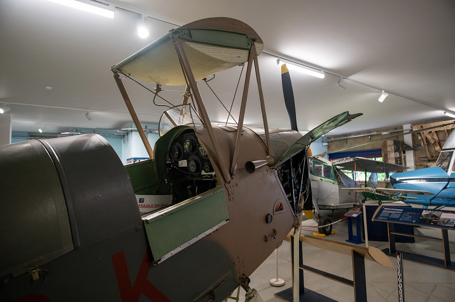 The image depicts an indoor museum exhibit featuring several vintage aircraft. The primary focus is on a fragile, old biplane with a visible cockpit and engine. Other aircraft are visible in the background, including a blue and white plane. Informational displays and barriers are set up around the exhibit area.
