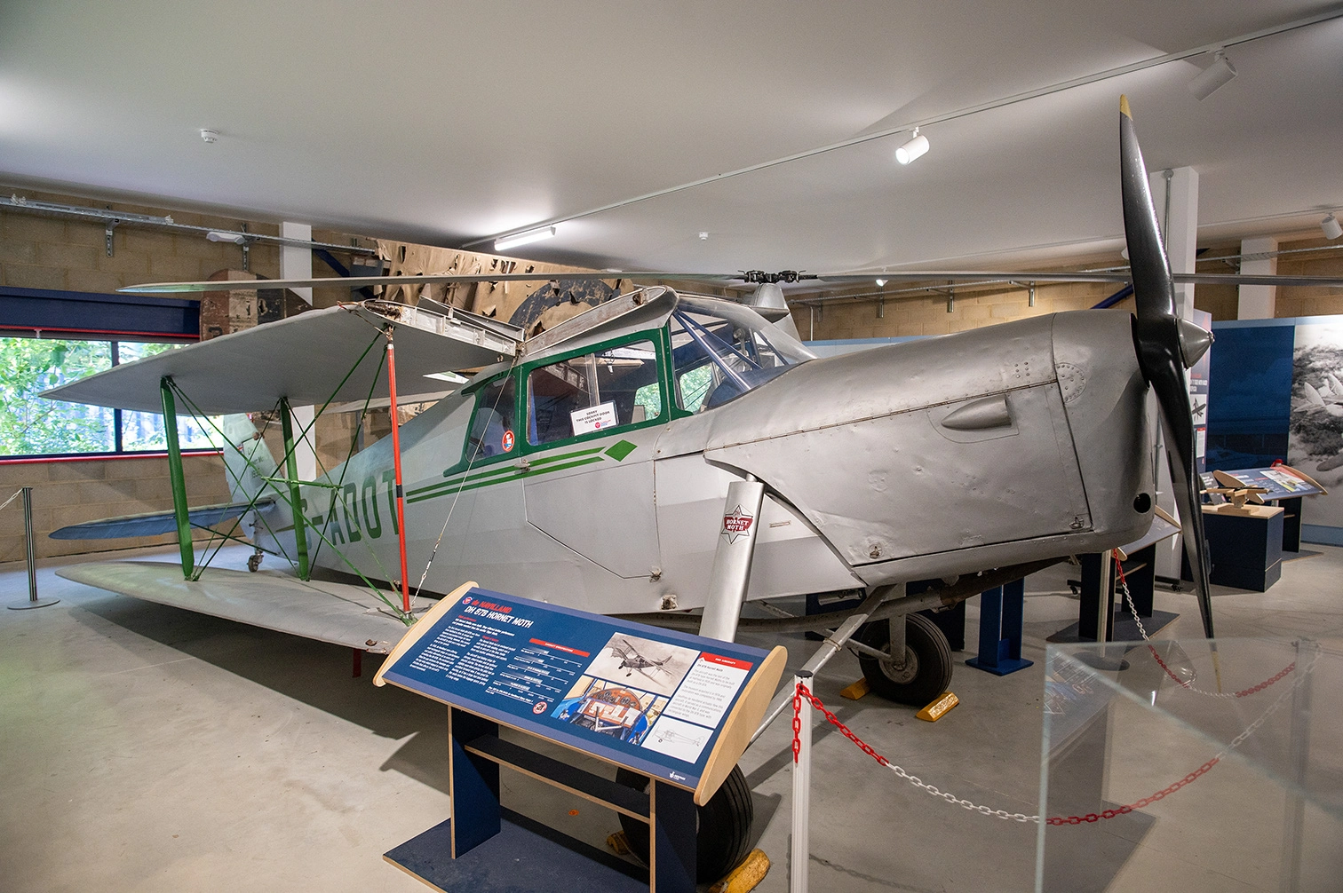 The image depicts a vintage aircraft displayed in a museum setting. The aircraft is a single-engine, single-wing plane with a propeller at the front. It is mounted on supports and surrounded by informational displays and barriers. The plane has a metallic body with green and white markings. The setting appears to be indoors with good lighting and additional exhibits in the background.