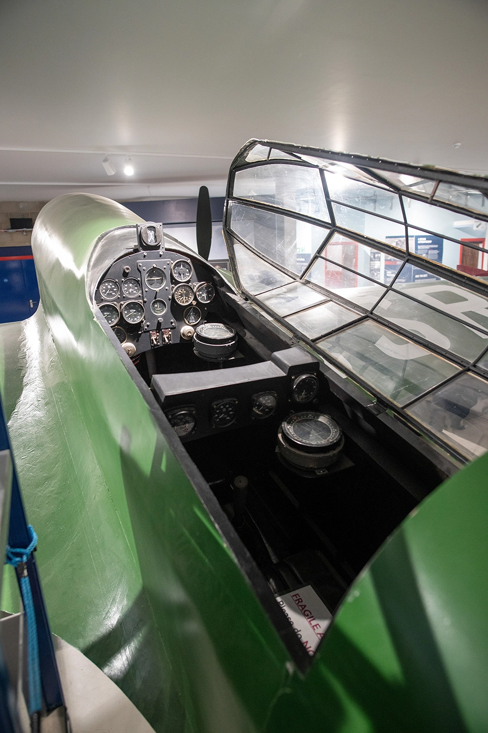 The image depicts the cockpit of a vintage aircraft, showcasing its control panel and instrumentation. The aircraft has a green exterior and a transparent canopy. The cockpit is equipped with various gauges, dials, and a control stick. The setting appears to be indoors, likely in a museum or exhibition space.