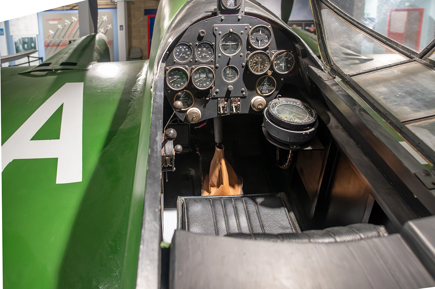 The image depicts the cockpit of a vintage military aircraft. The interior features an array of analog dials and gauges on the instrument panel, a control stick with a leather cover, and a throttle control. The aircraft's exterior is painted in a green color, with a large white number '4' visible on the side. The cockpit appears to be part of a museum exhibit, showcasing historical aviation equipment.